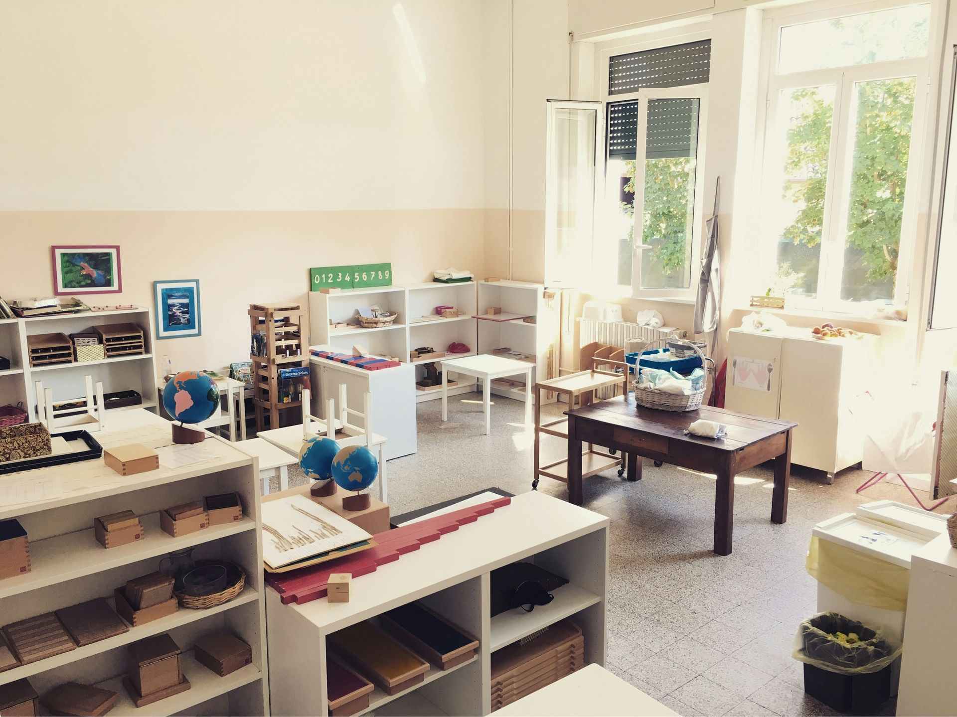 A well-lit Montessori classroom with white shelves, various learning materials, and natural light from large windows.