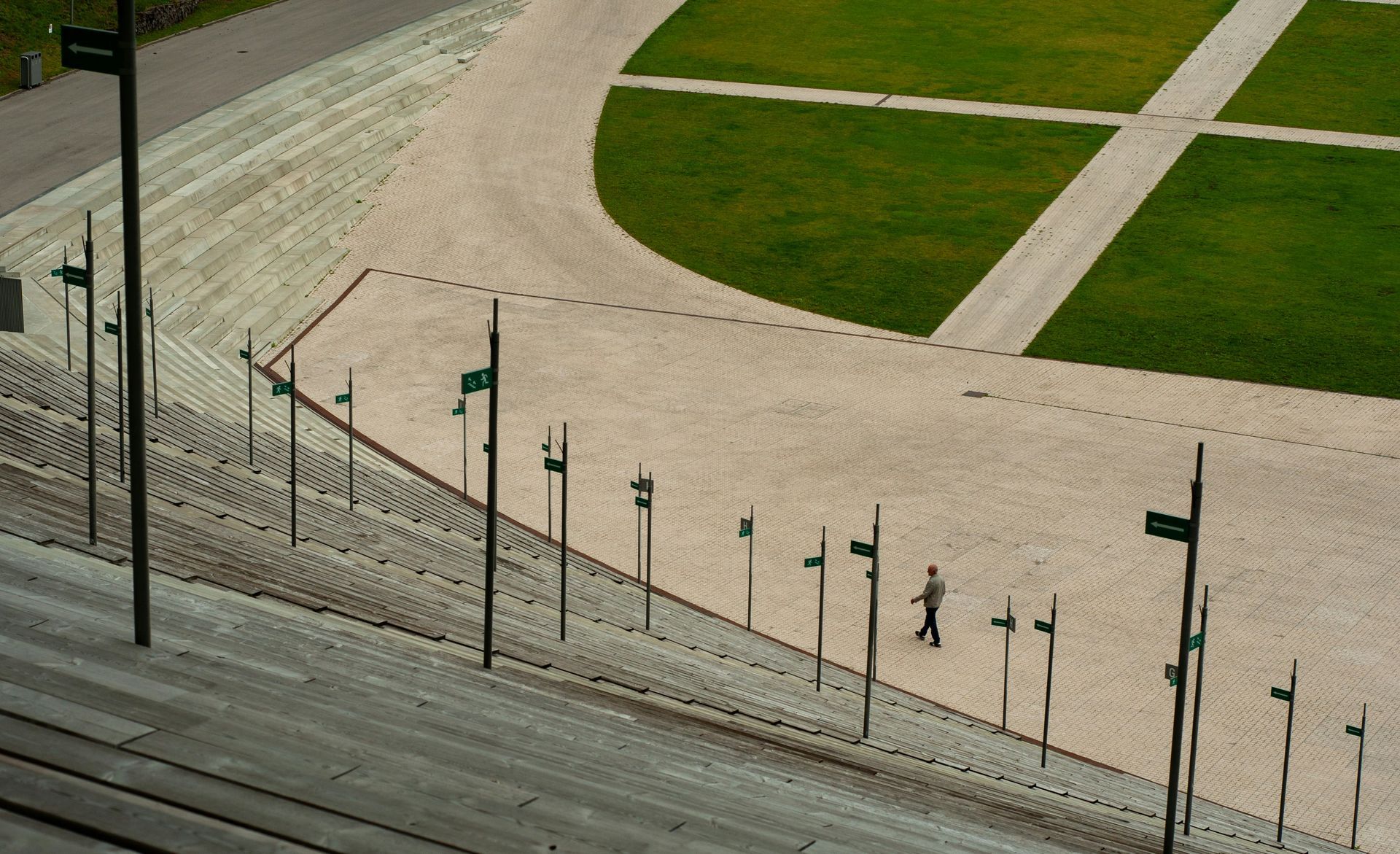 View from above of a large, stepped concrete structure with a pathway and grassy areas; person walking.