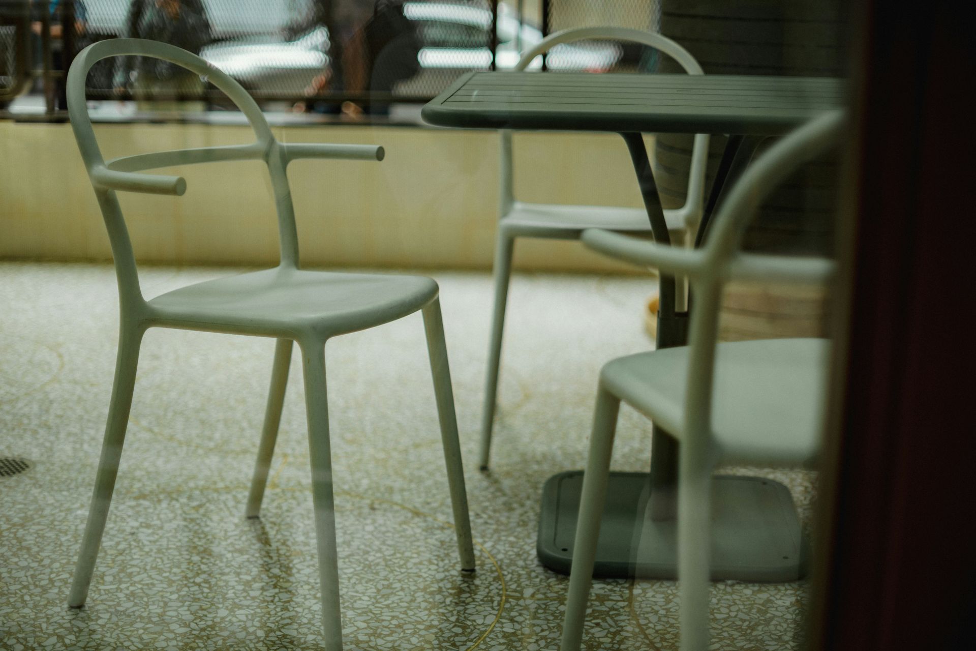 Three white chairs sit at a table outside a cafe, viewed through a window on a rainy day.
