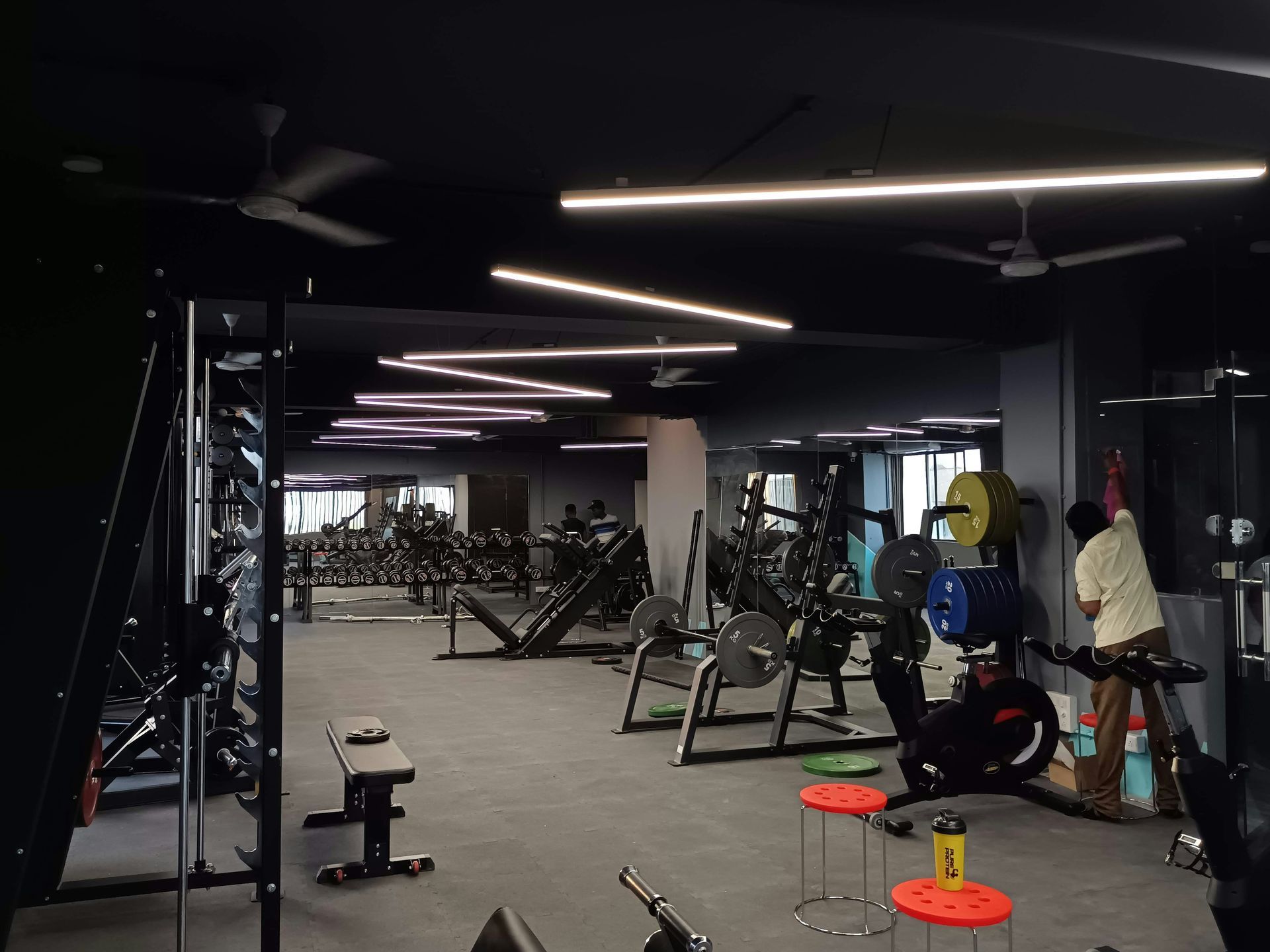 Gym interior with weightlifting equipment. A person stands near a weight machine. Dark walls, overhead lights.