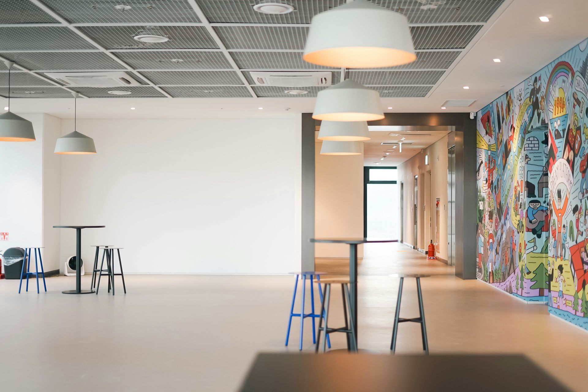 Bright, open room with tables and stools, plus colorful mural on a wall; pendant lights hang from patterned ceiling.