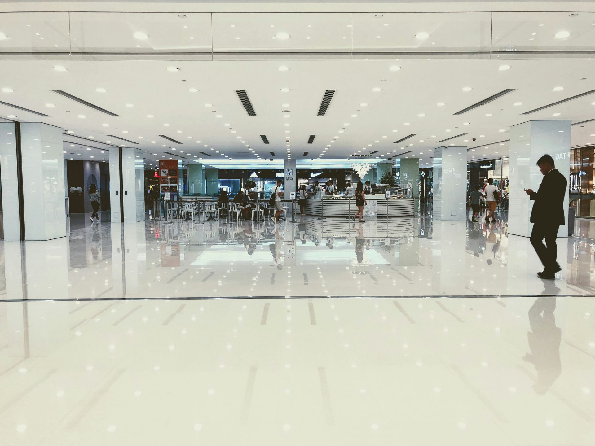 A person in a black coat stands in a brightly lit shopping mall with reflective floors.