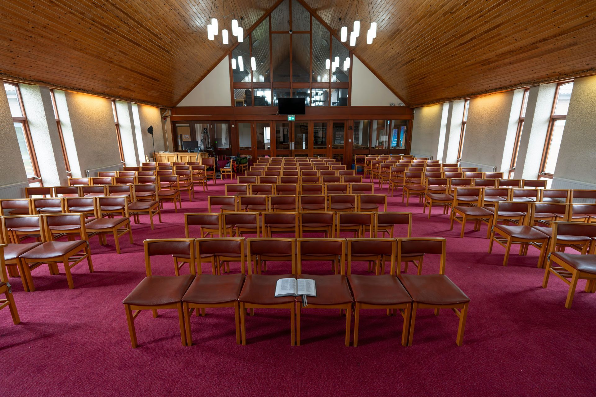 Rows of empty wooden chairs in a church auditorium, with a book on a front row. Red carpet and wood paneling.