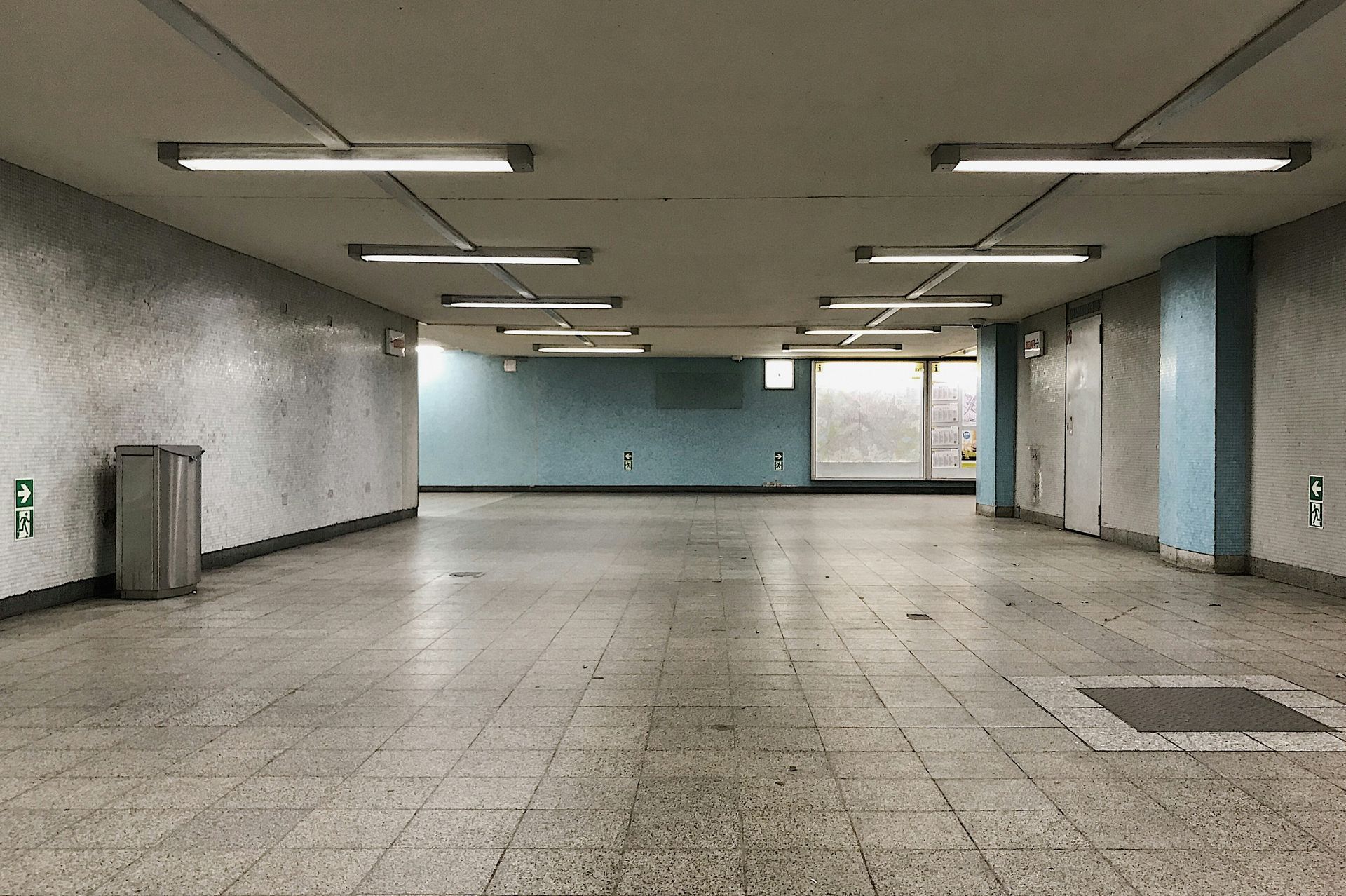 Empty, tiled, underground pedestrian tunnel with bright fluorescent lights; blue and white walls; trash bin on left.