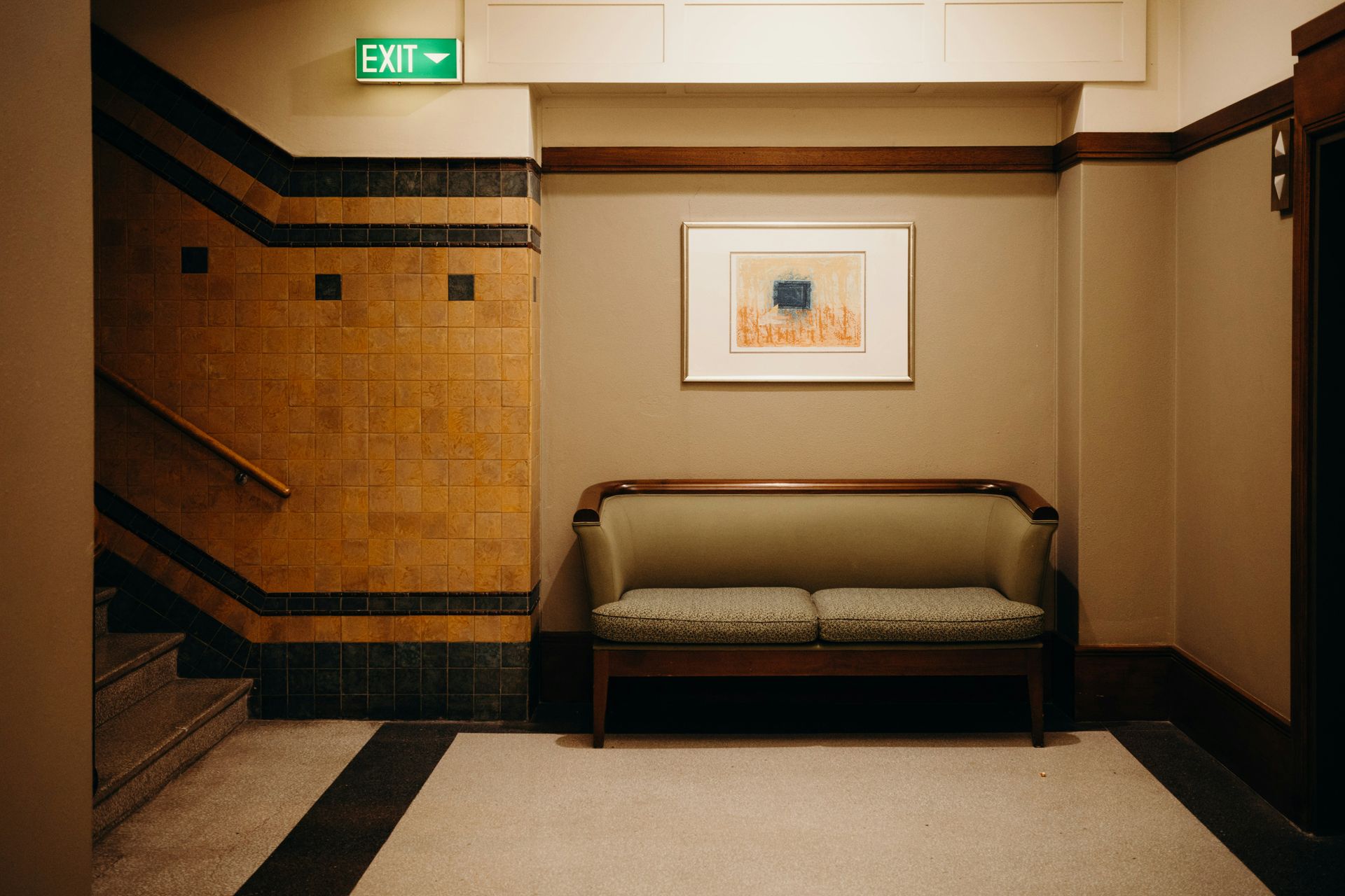 Hallway with stairs, a bench, artwork, and an exit sign, with tan walls and patterned tile.