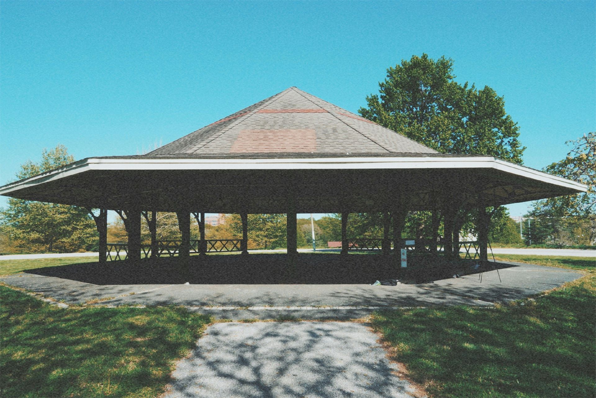 Pavilion with angled roof, supported by columns. Located in a park with grass and trees under a blue sky.