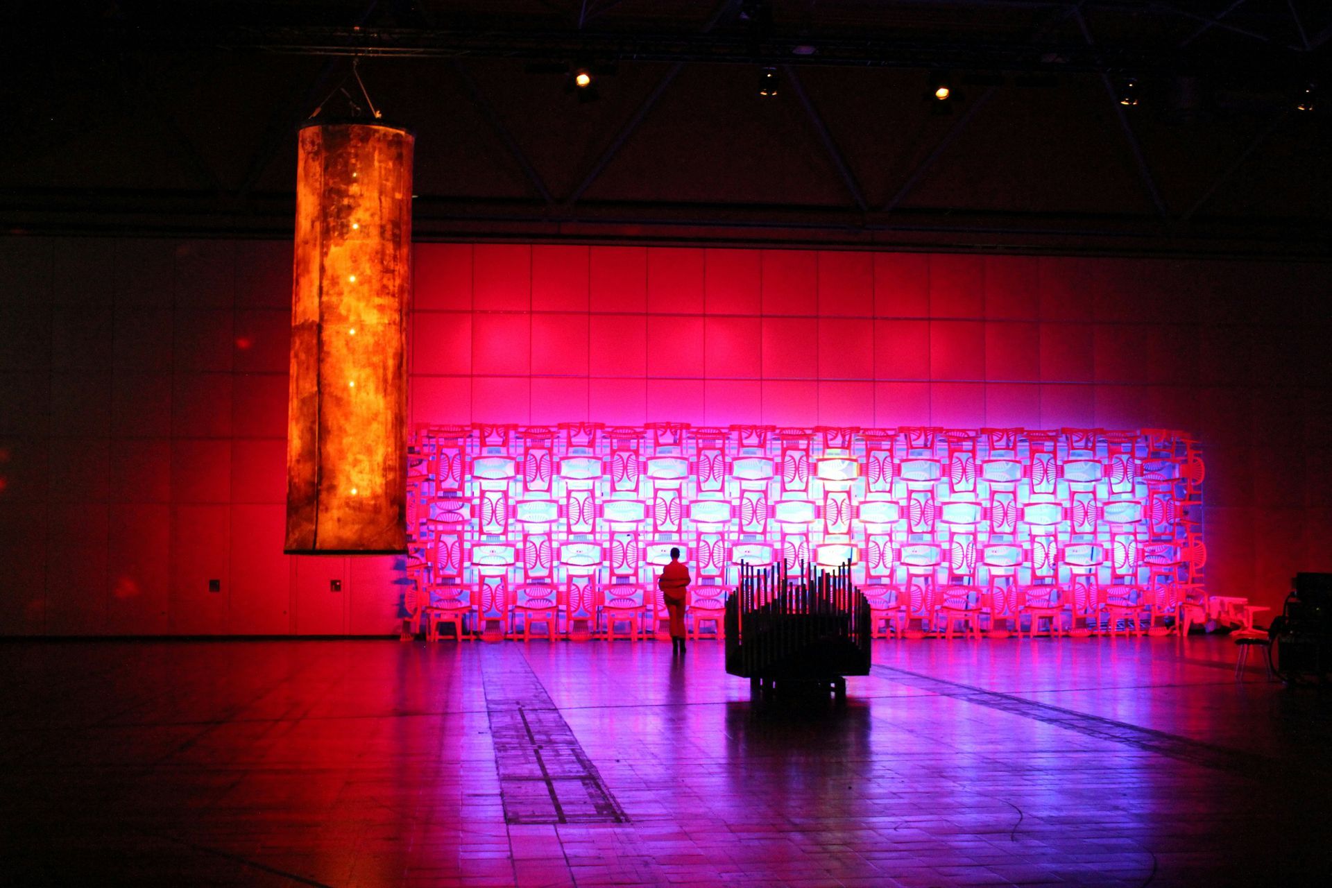 Red-lit art installation: a rectangular light display, hanging cylinder, and person in the center.