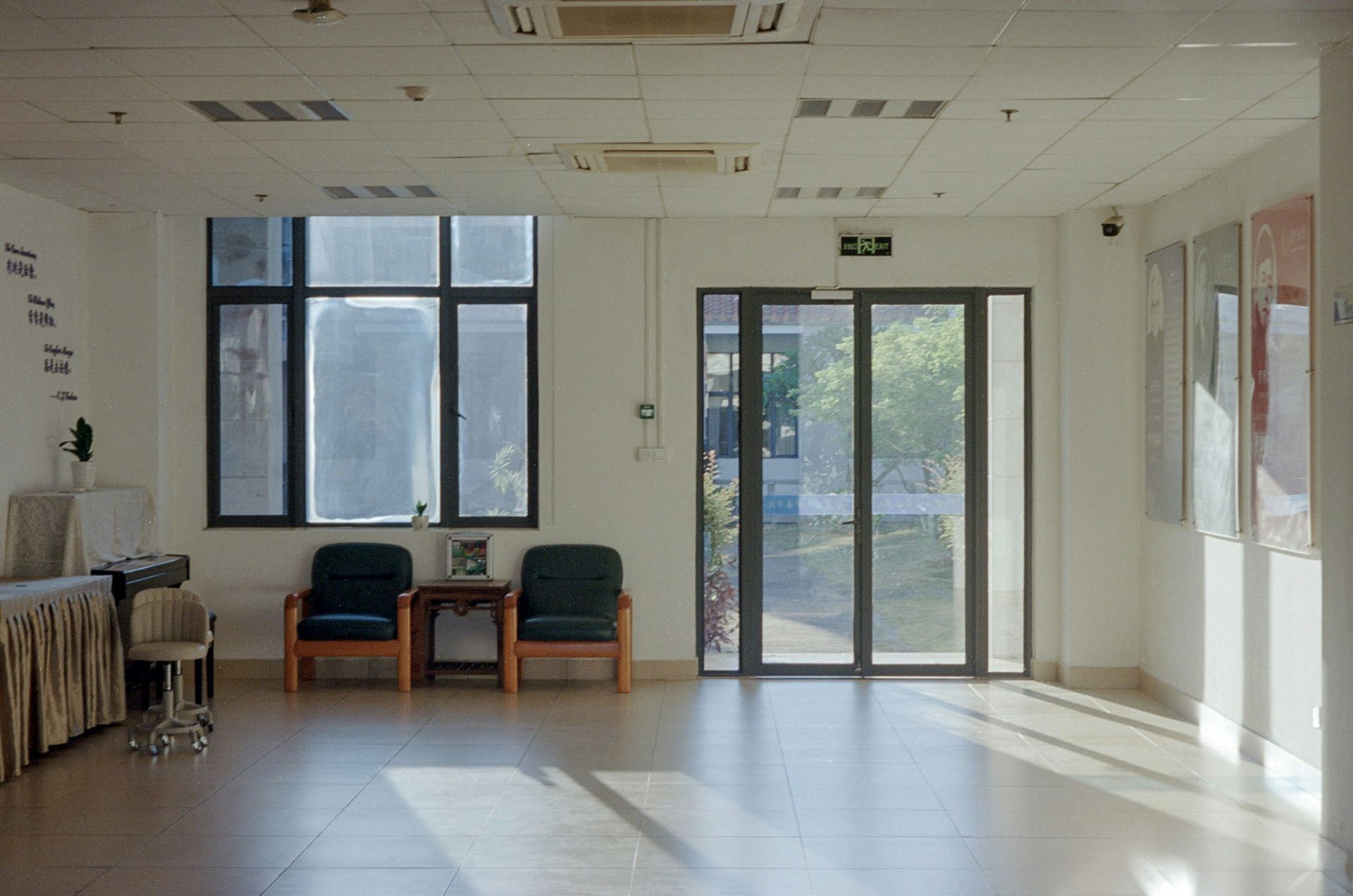 Empty lobby with large windows, sliding glass doors, chairs, and table. Sunlight streams in.