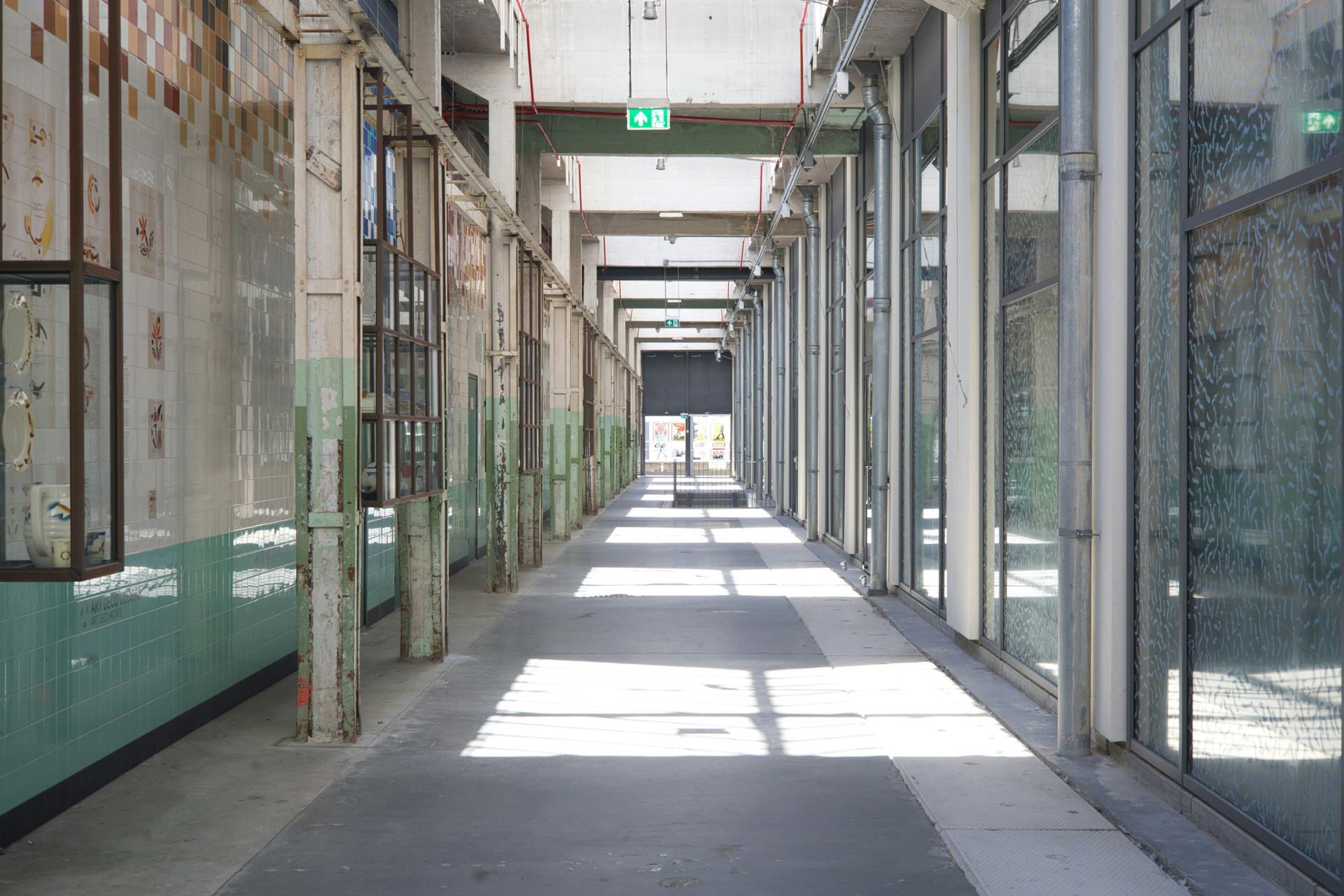 Long hallway with concrete floor and pillars. Sunlight streams through glass windows, illuminating the space.