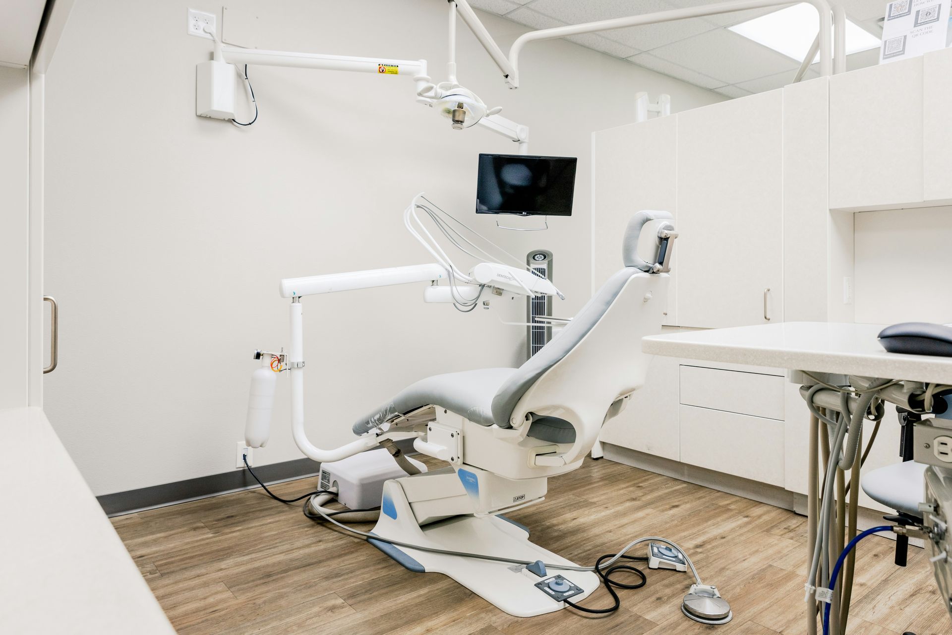 Dental office with white chair, tools, and cabinets; brown floor; and a small tv.