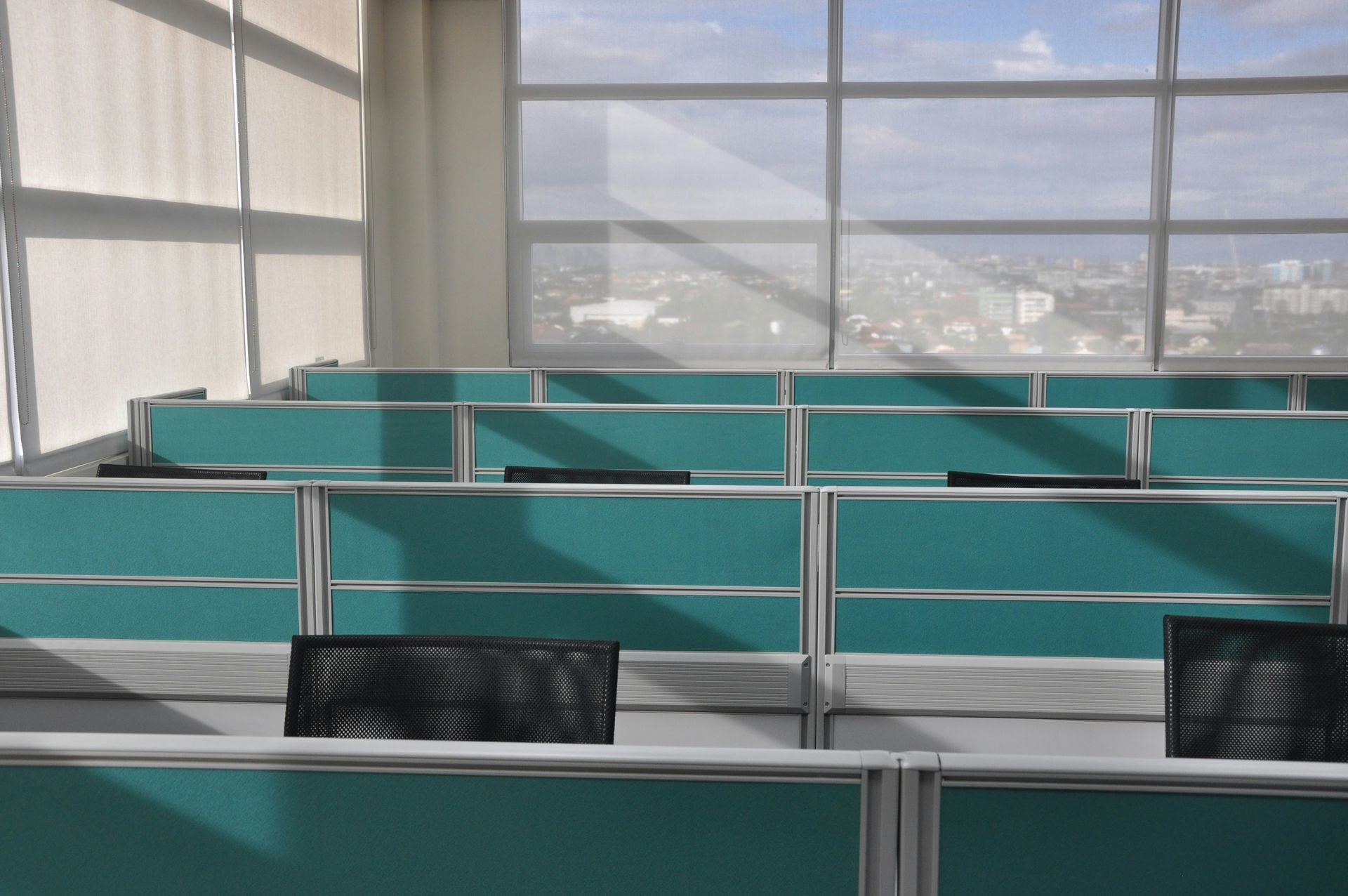 Rows of teal cubicles in a brightly lit office with a large window overlooking a cityscape.