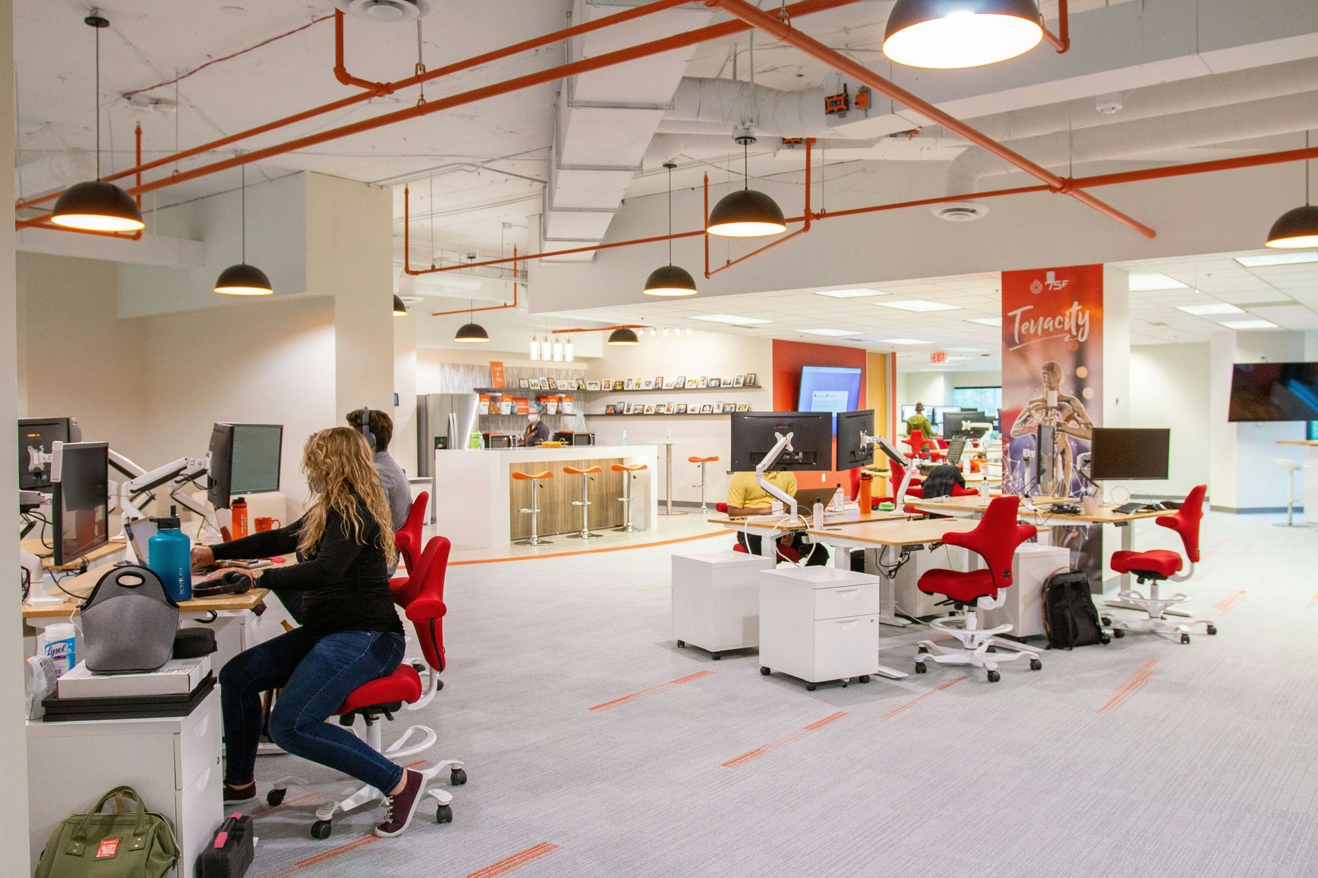 Open office with people working at desks, red chairs, and overhead lighting.