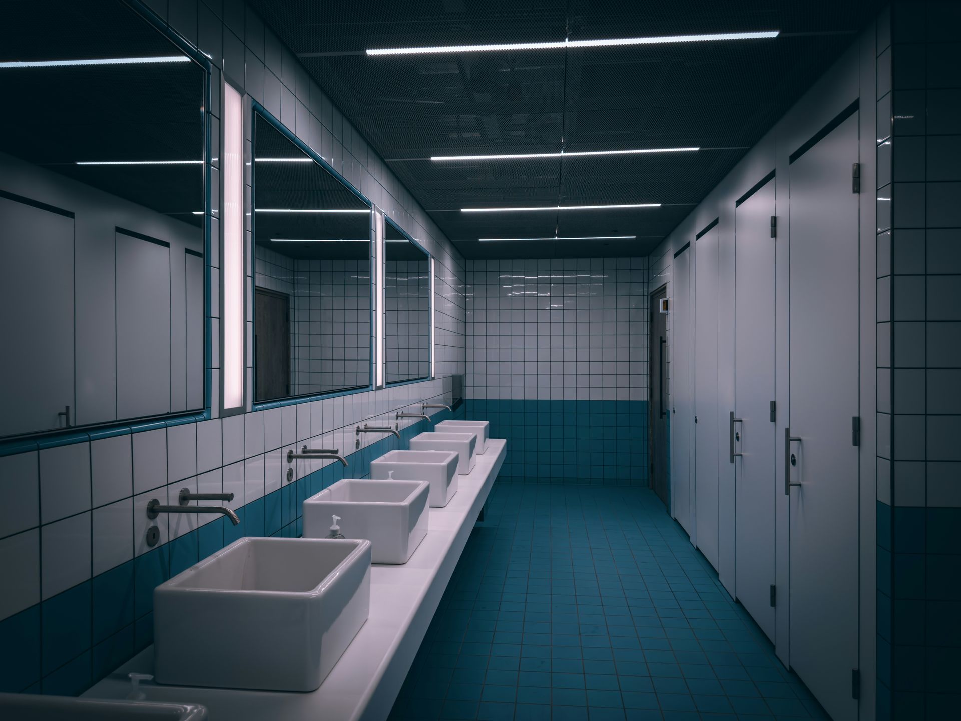Public restroom with white sinks, mirrors, and stalls, blue tiled floor and walls, overhead lights.