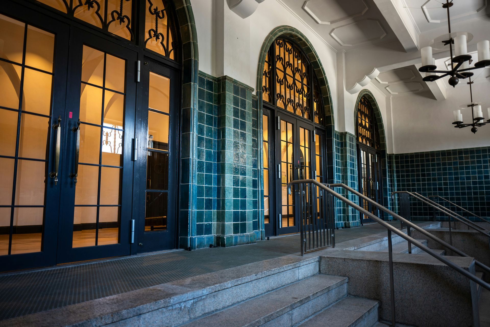 Hallway with blue-tiled walls, arched glass doors, and steps leading up. Black railings and chandeliers.