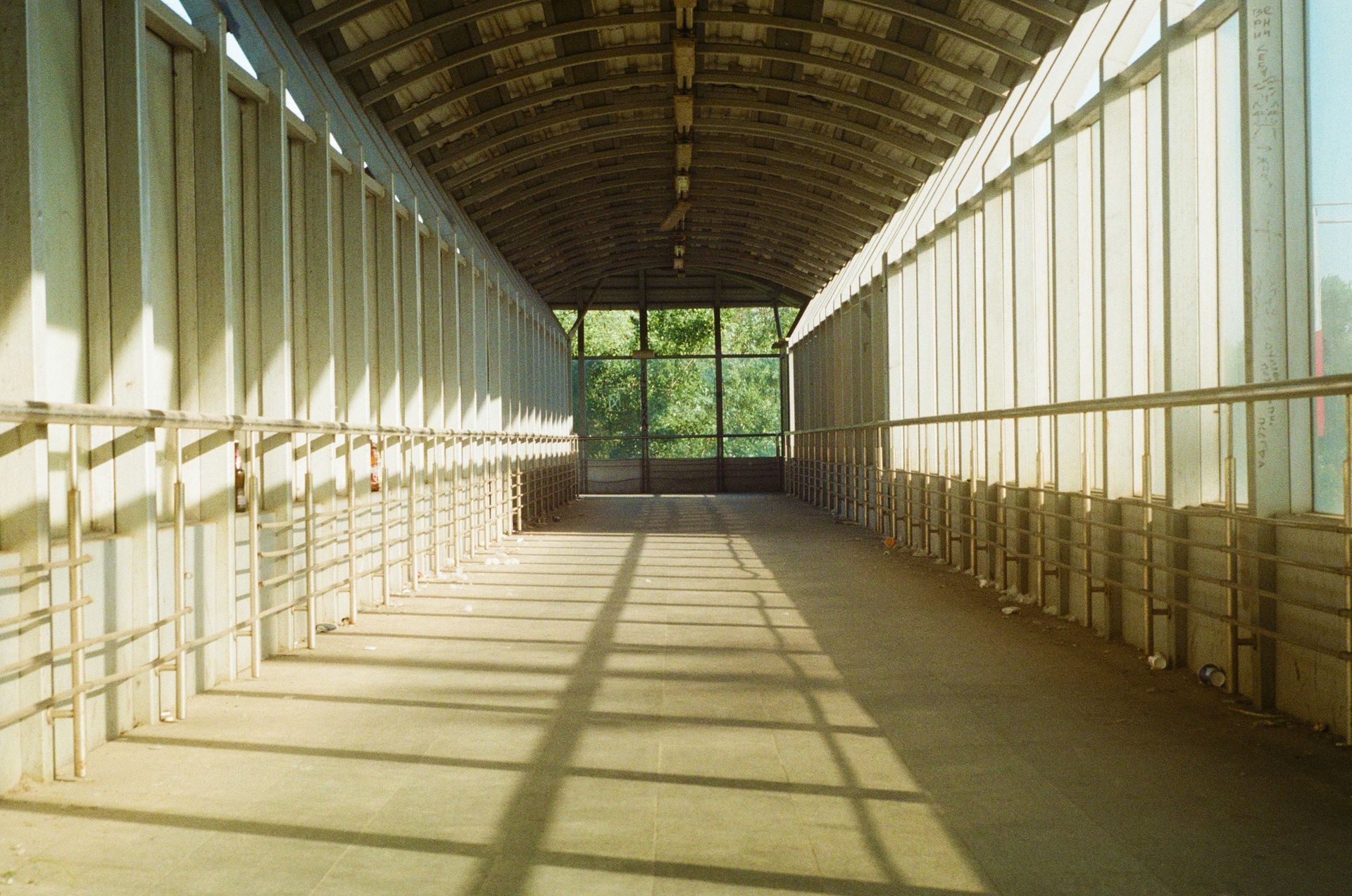 Overhead walkway. Arched roof, walls with vertical panels, sunlight casts shadows. View to green trees.