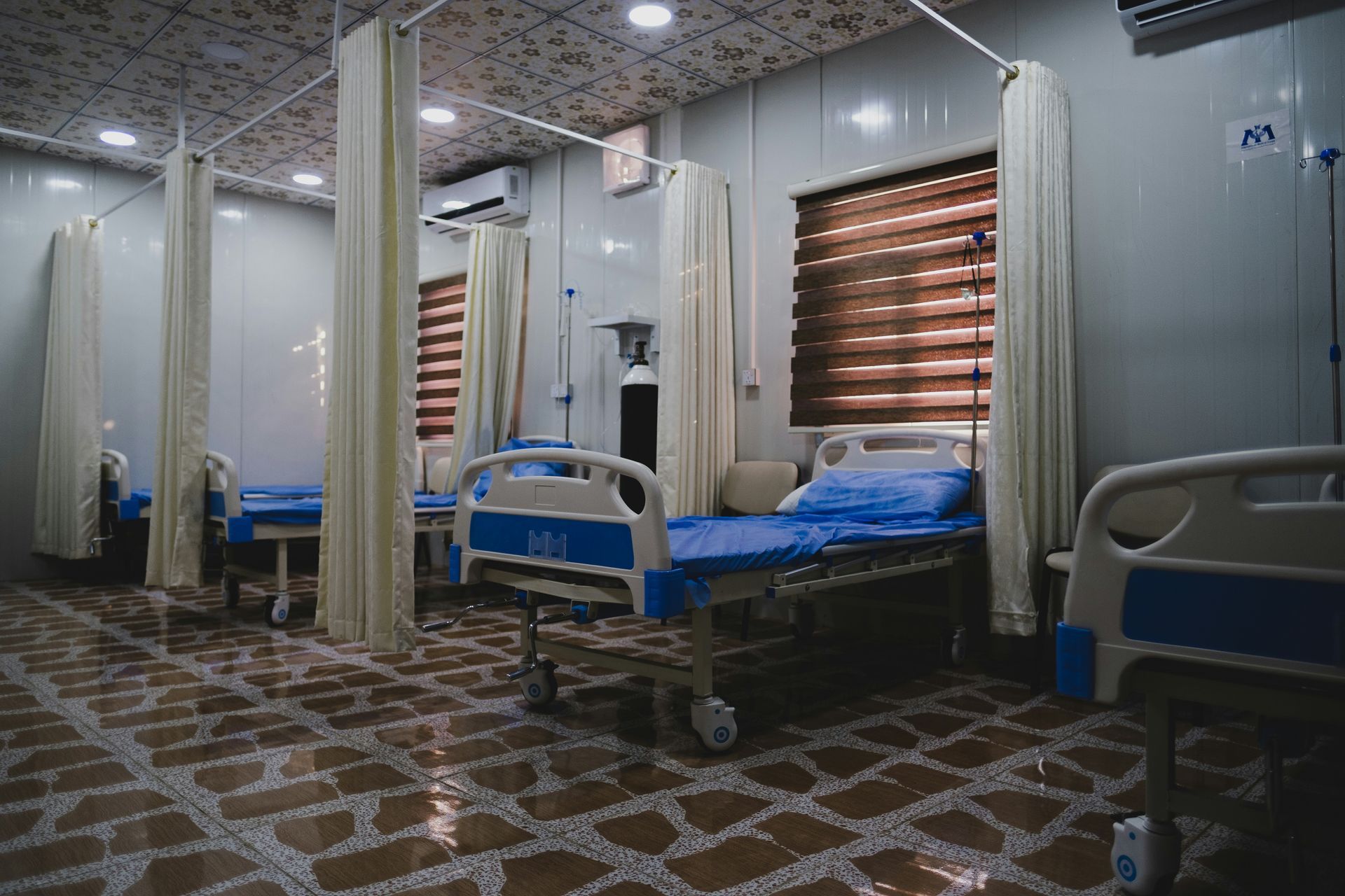 Hospital room with beds, curtains, and tiled floor. Windows with blinds and oxygen tank visible.