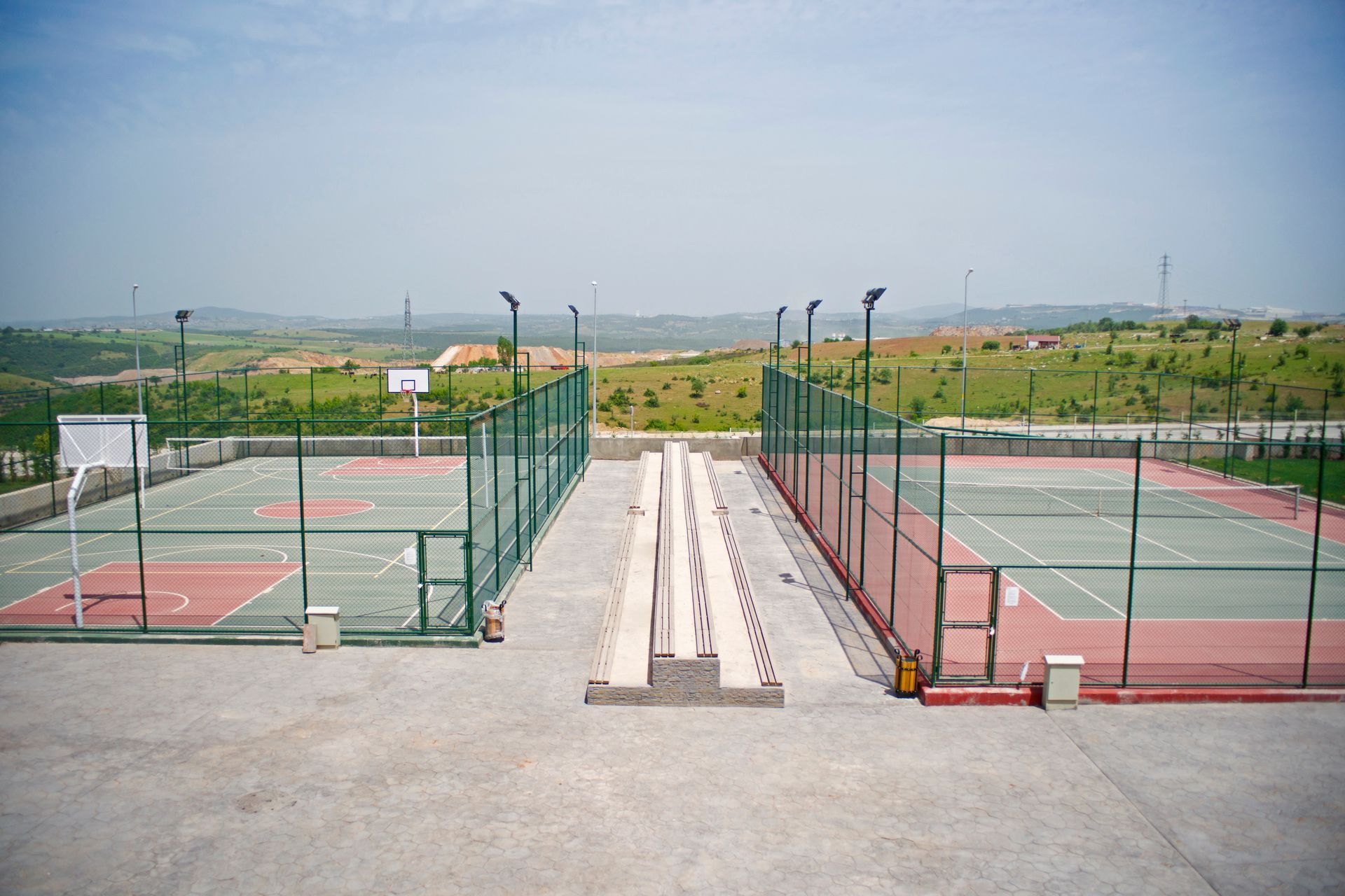 Two outdoor sports courts separated by a central walkway, with a landscape background.