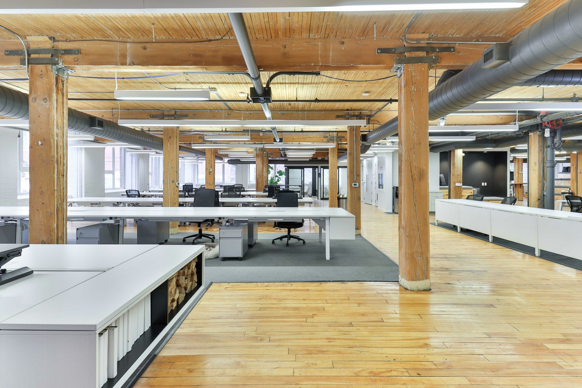 Bright, open-plan office with wooden beams and columns. White desks and cabinets fill the space, natural light.