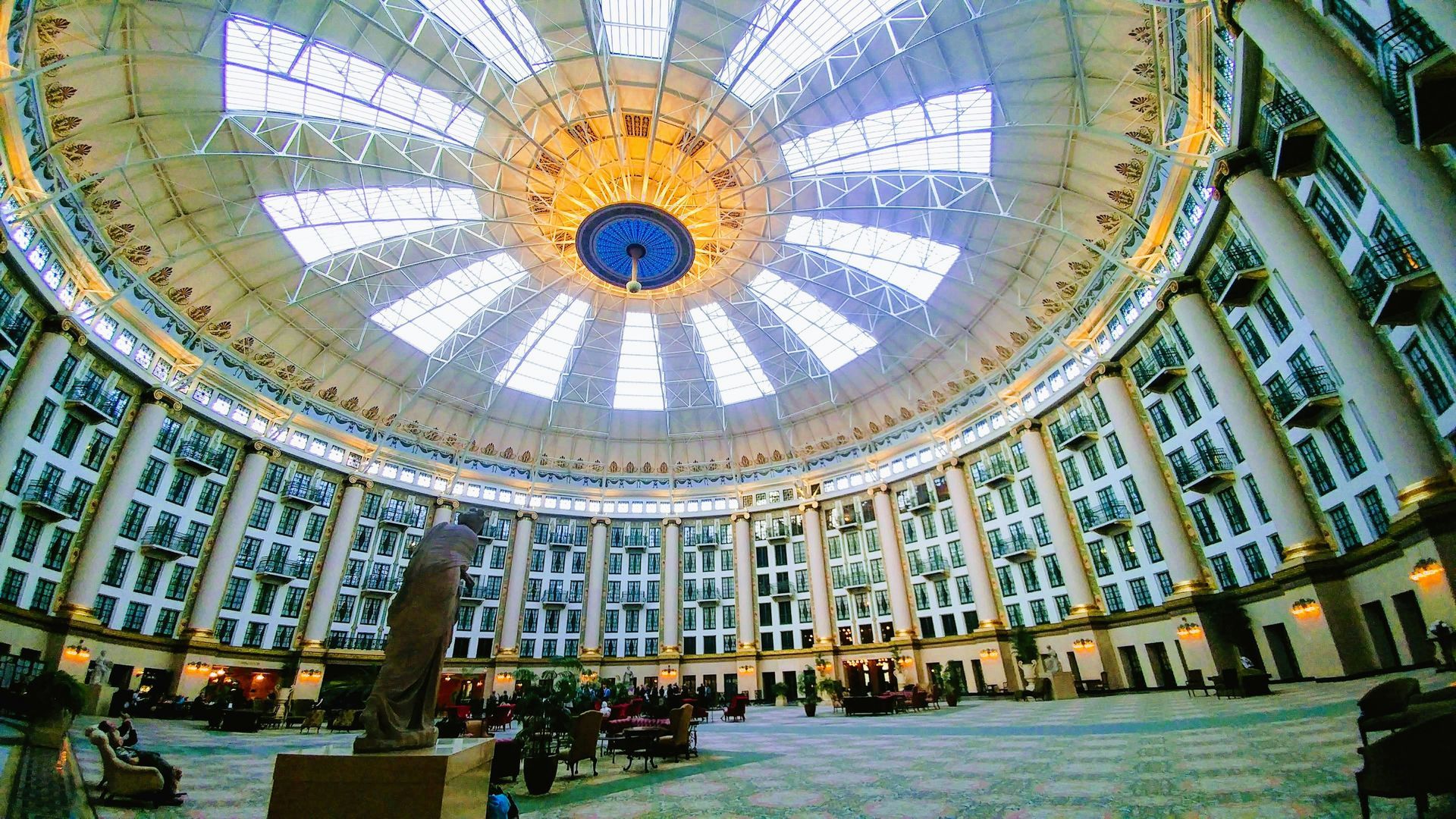 Interior of building with large domed glass ceiling, columns, statues, and many people.