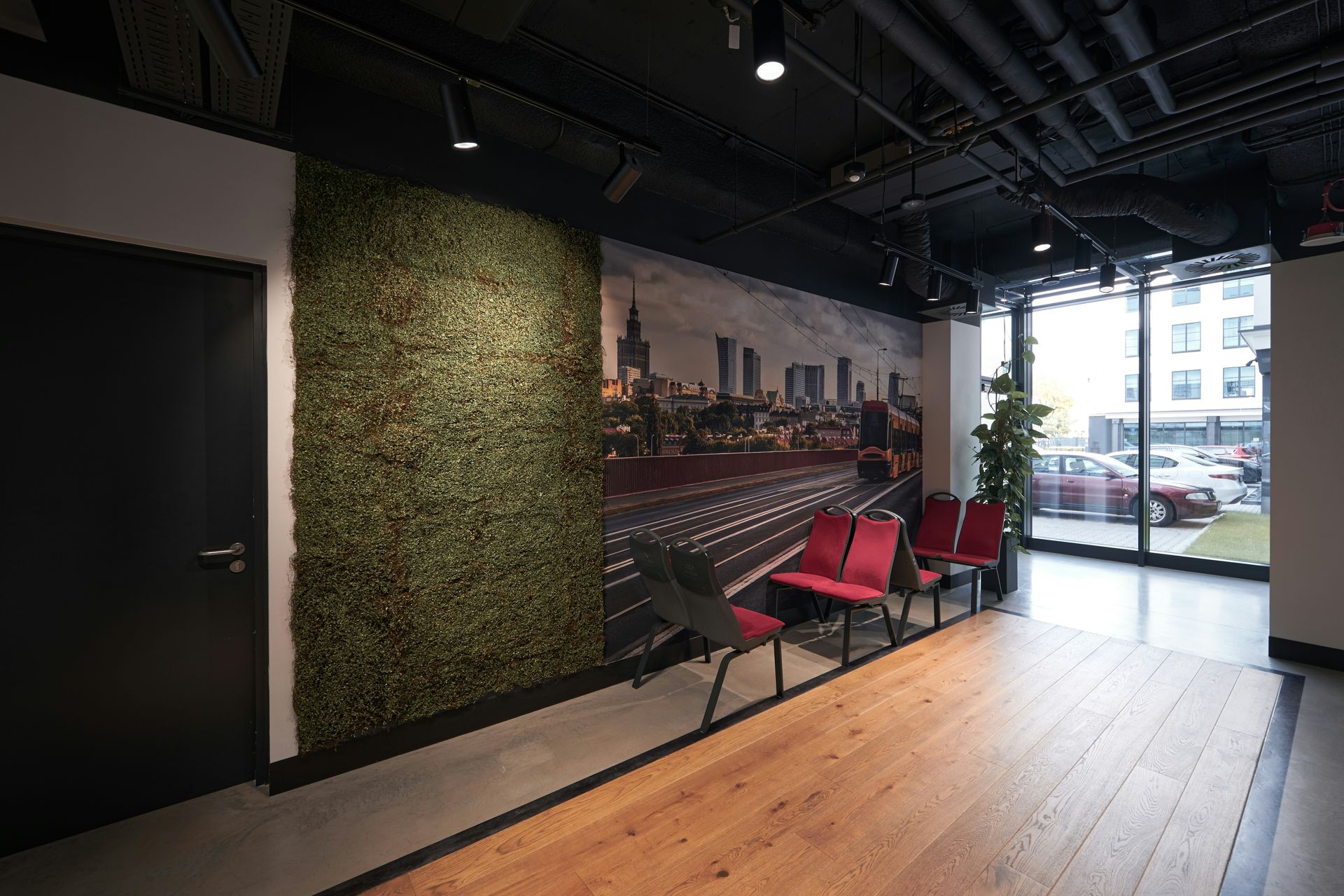 A modern waiting area with a green wall, cityscape mural, red chairs, and a black door next to a window.