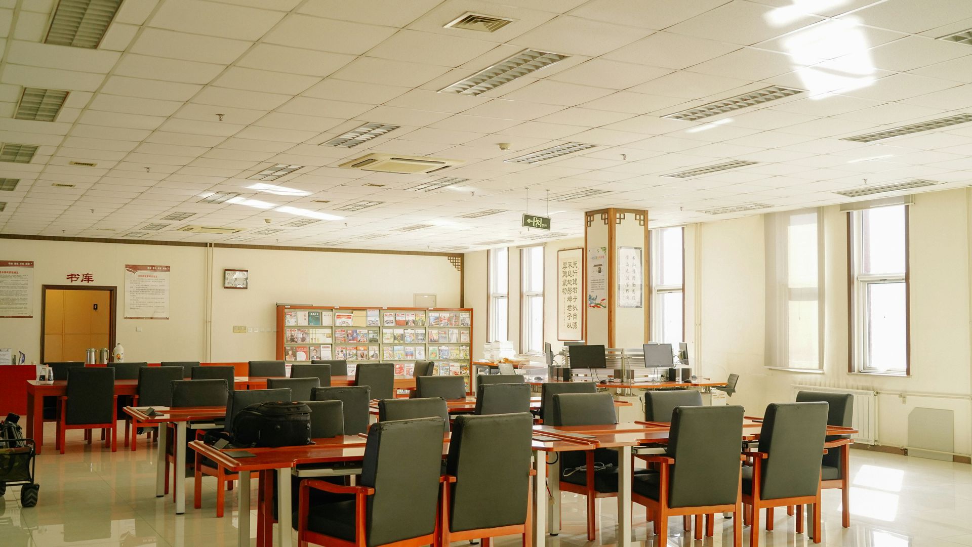 Interior of a library with tables, chairs, bookshelves, and computers; a bright ceiling with recessed lights.