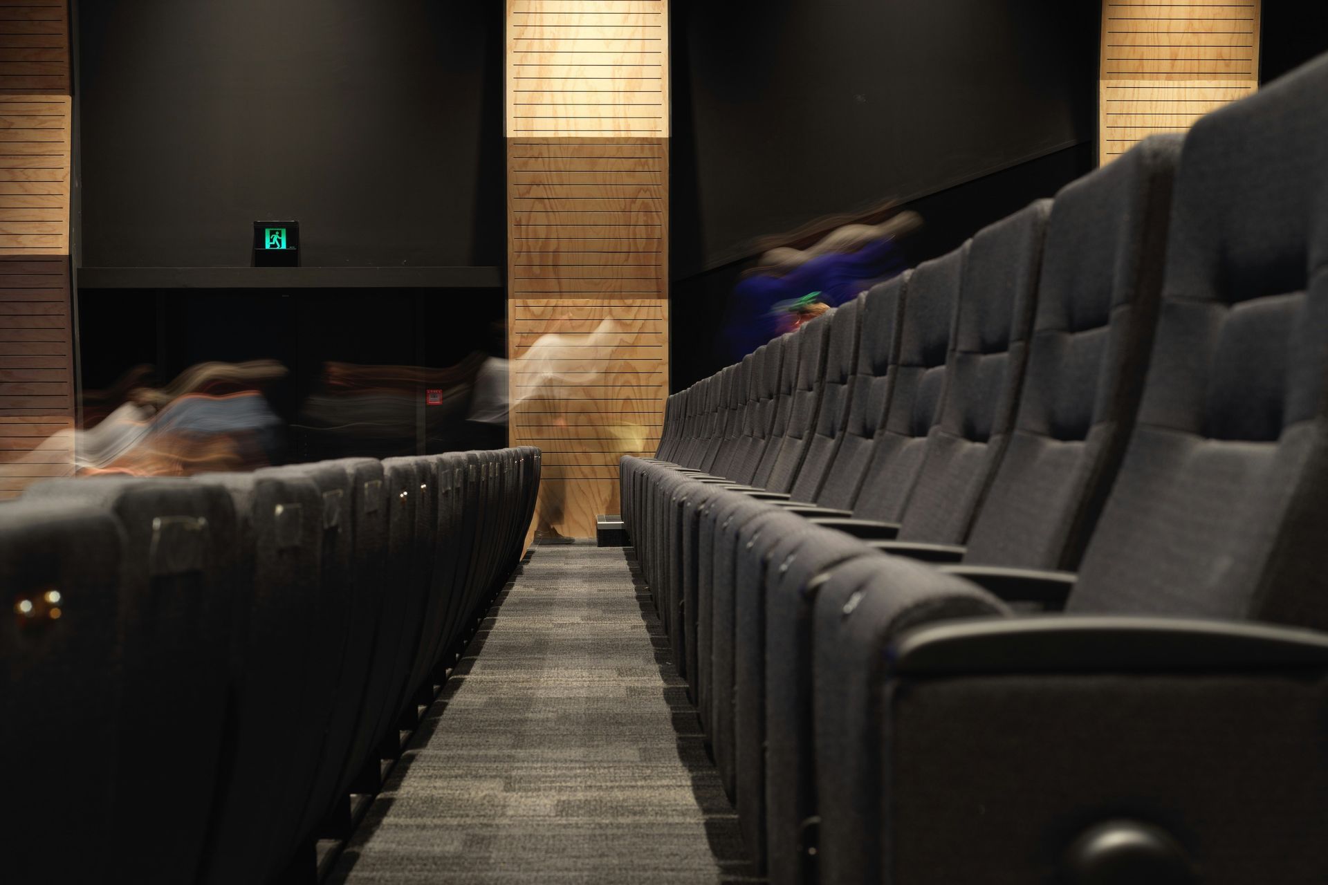 Rows of black theater seats in an empty auditorium. People walking past in the distance.