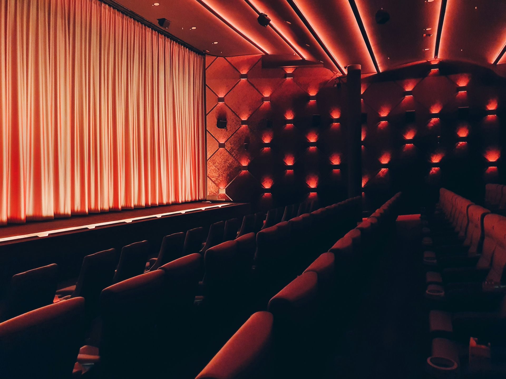 Red-lit cinema interior with rows of seats facing a screen hidden behind a curtain.