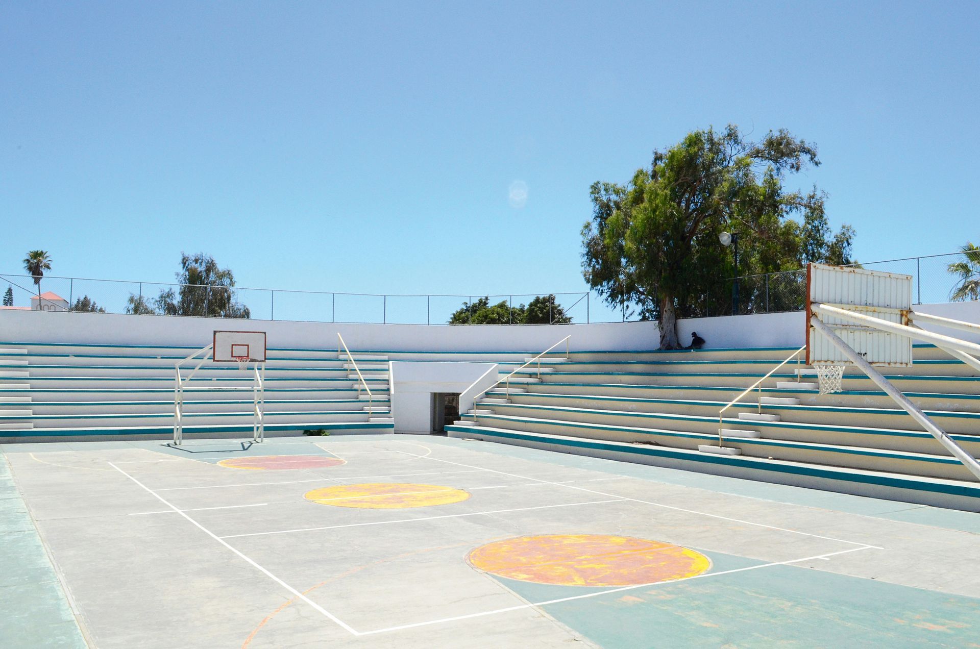 Outdoor concrete sports court with bleachers, basketball hoop, soccer goal, under a bright blue sky.