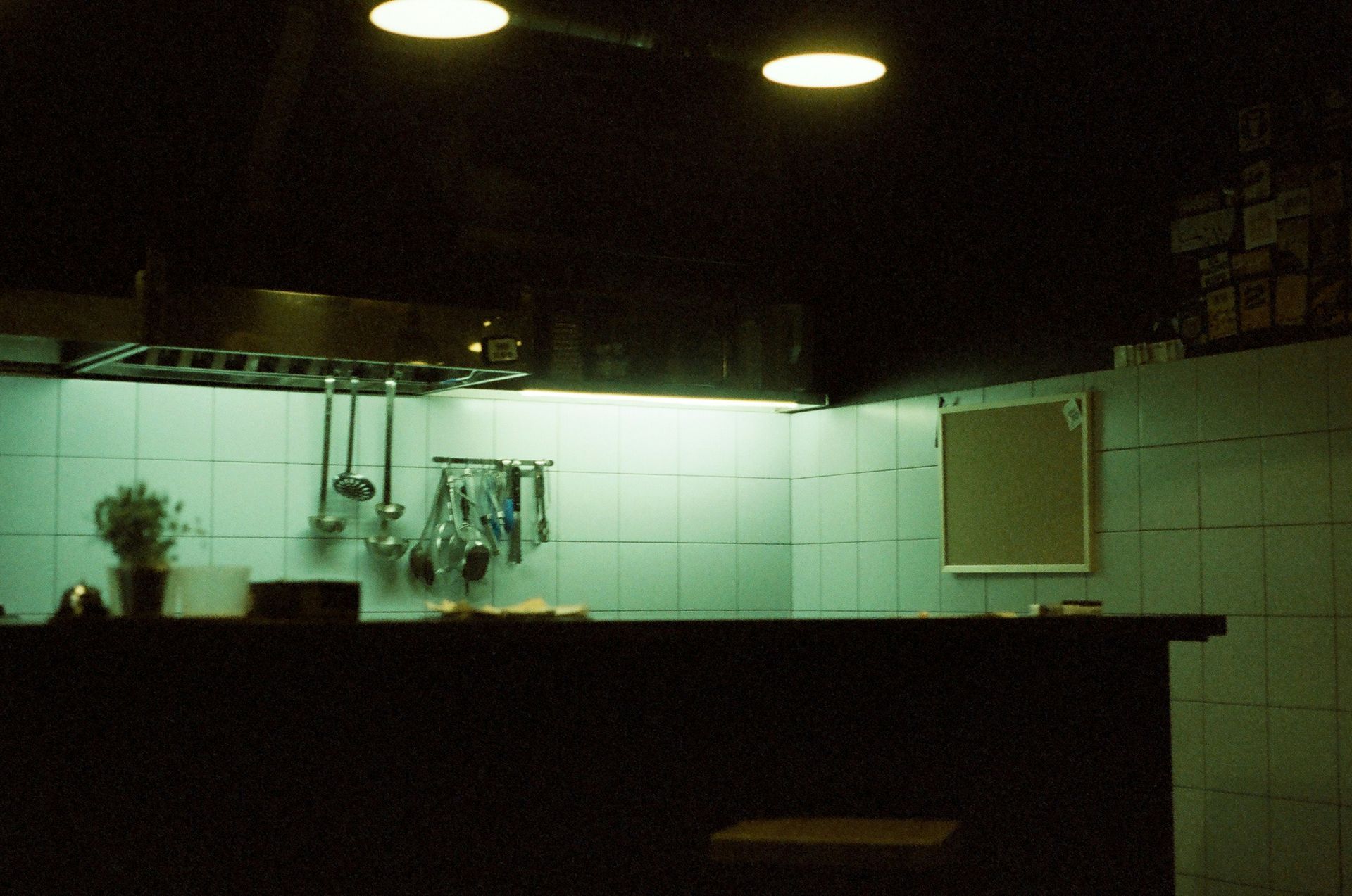 Dark restaurant kitchen. Green-tiled walls, stainless steel accents, and overhead lighting.