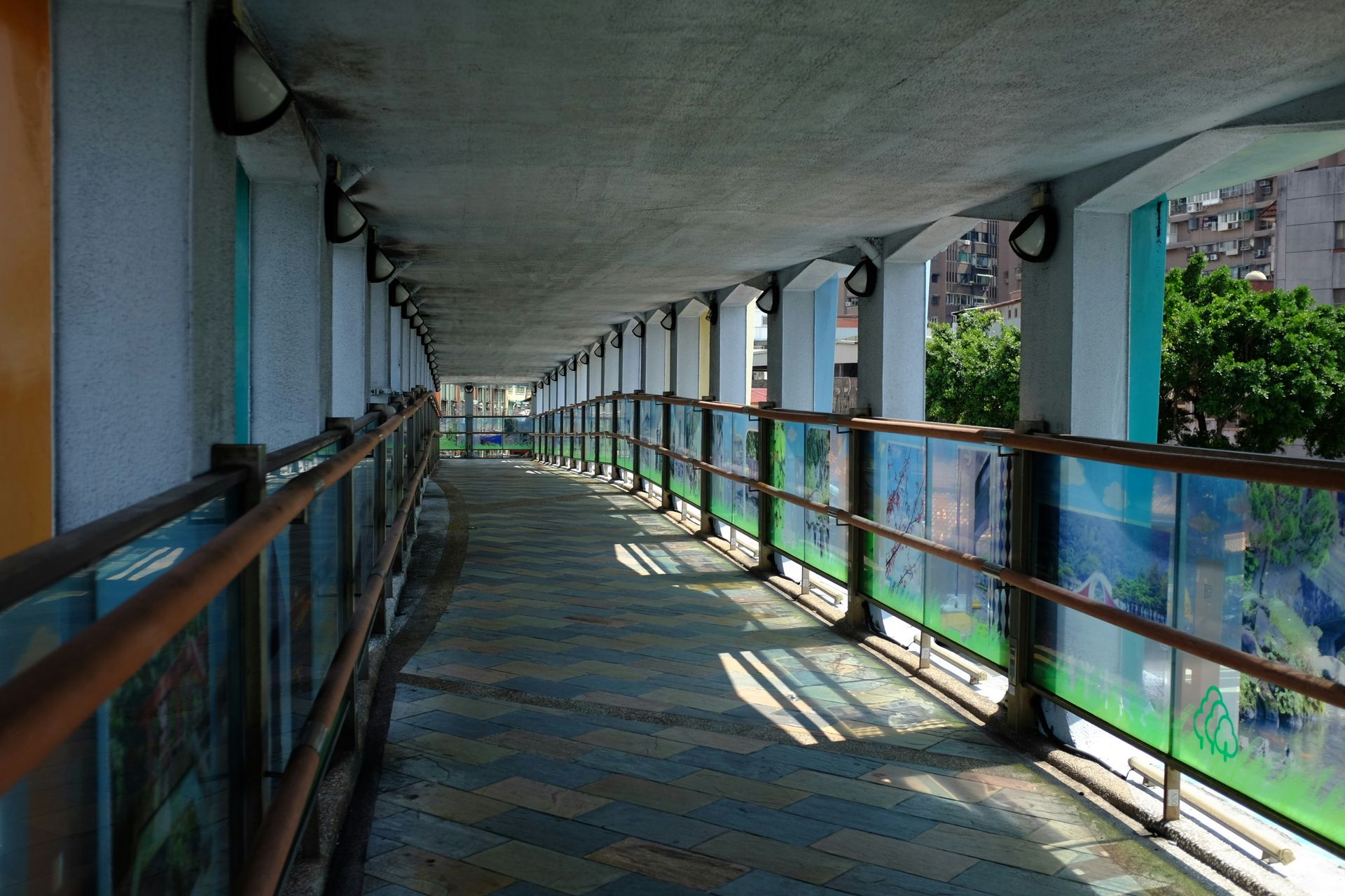 Covered walkway with glass panels and metal railings, concrete ceiling, sunlight casting shadows.