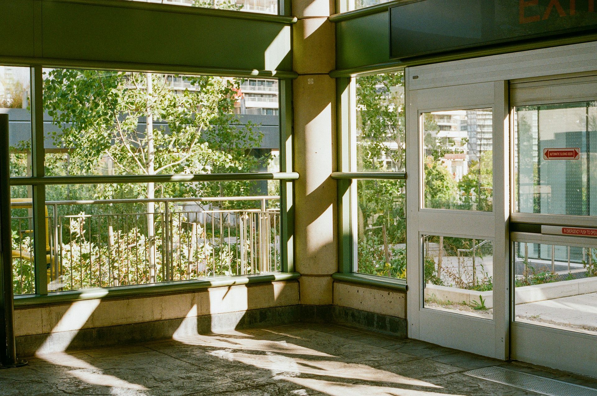 Interior of a building with sunlight. Windows reveal trees and a door on the right.