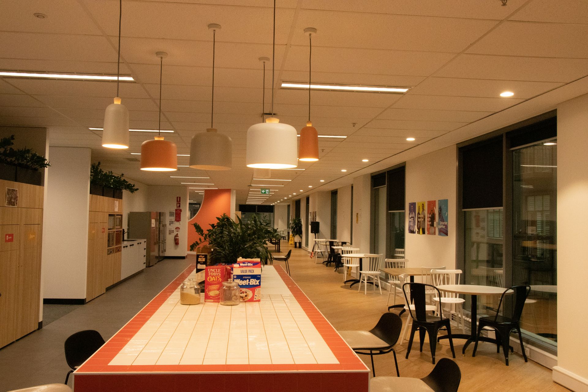 Modern office break room with long counter, tables, and pendant lights.
