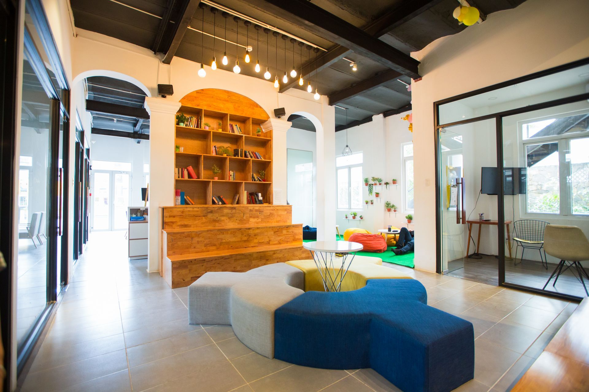 Lounge area with a colorful sectional sofa, bookshelves, and glass-walled rooms. Warm lighting and bright colors.