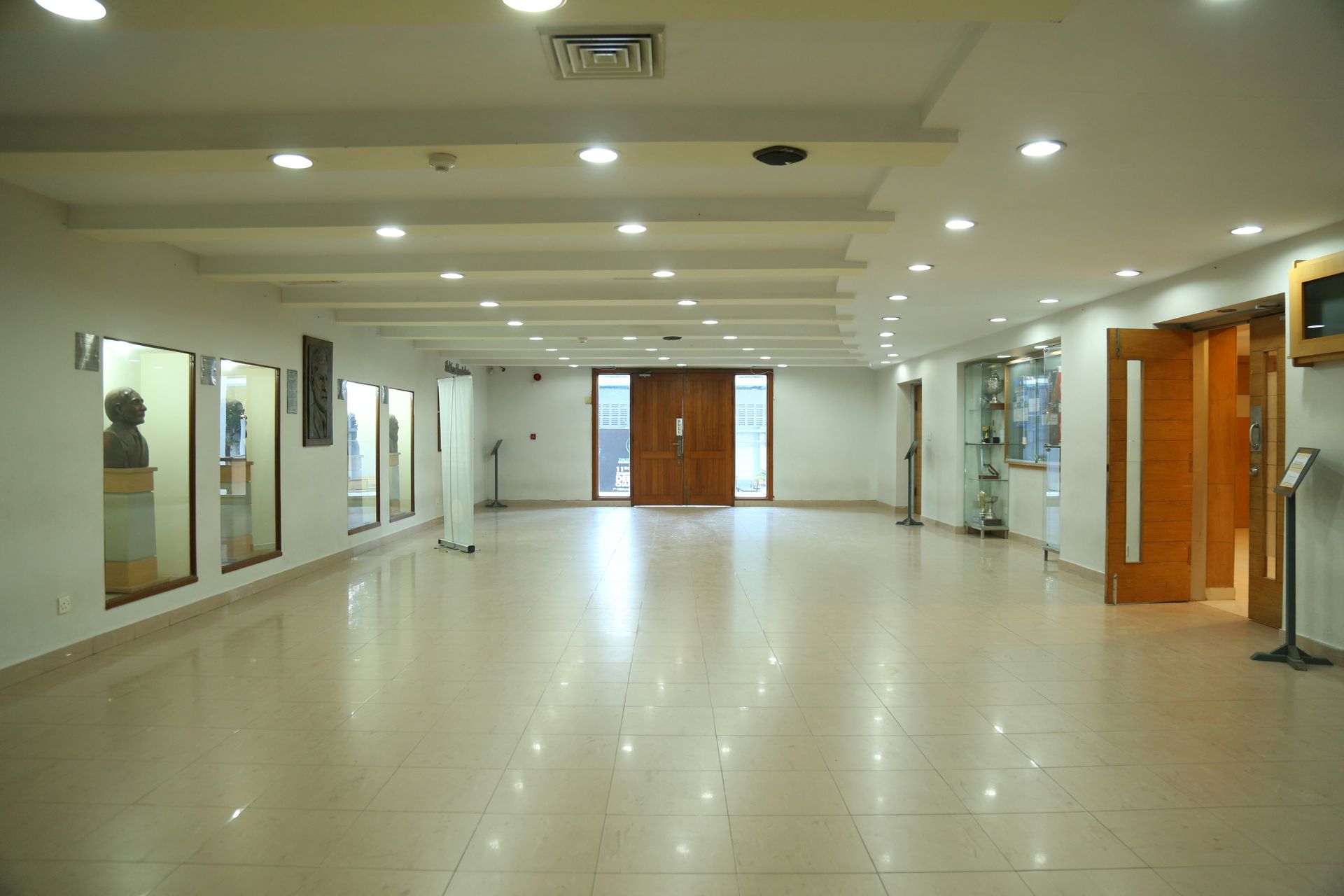 Hallway with display cases, wood doors, and overhead lighting. Light beige walls and floor.