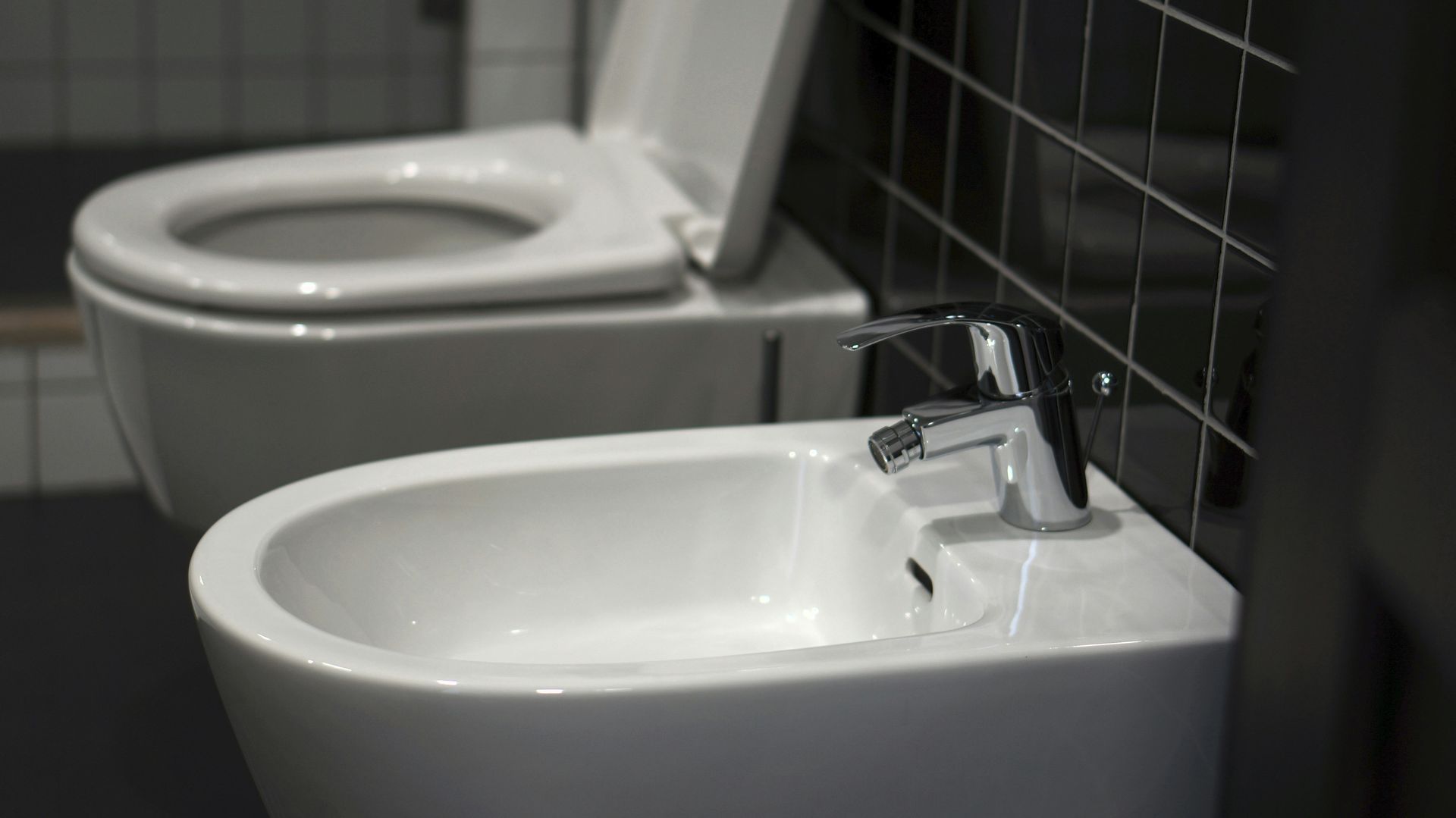 White bidet and toilet in a modern bathroom setting with dark tiles.