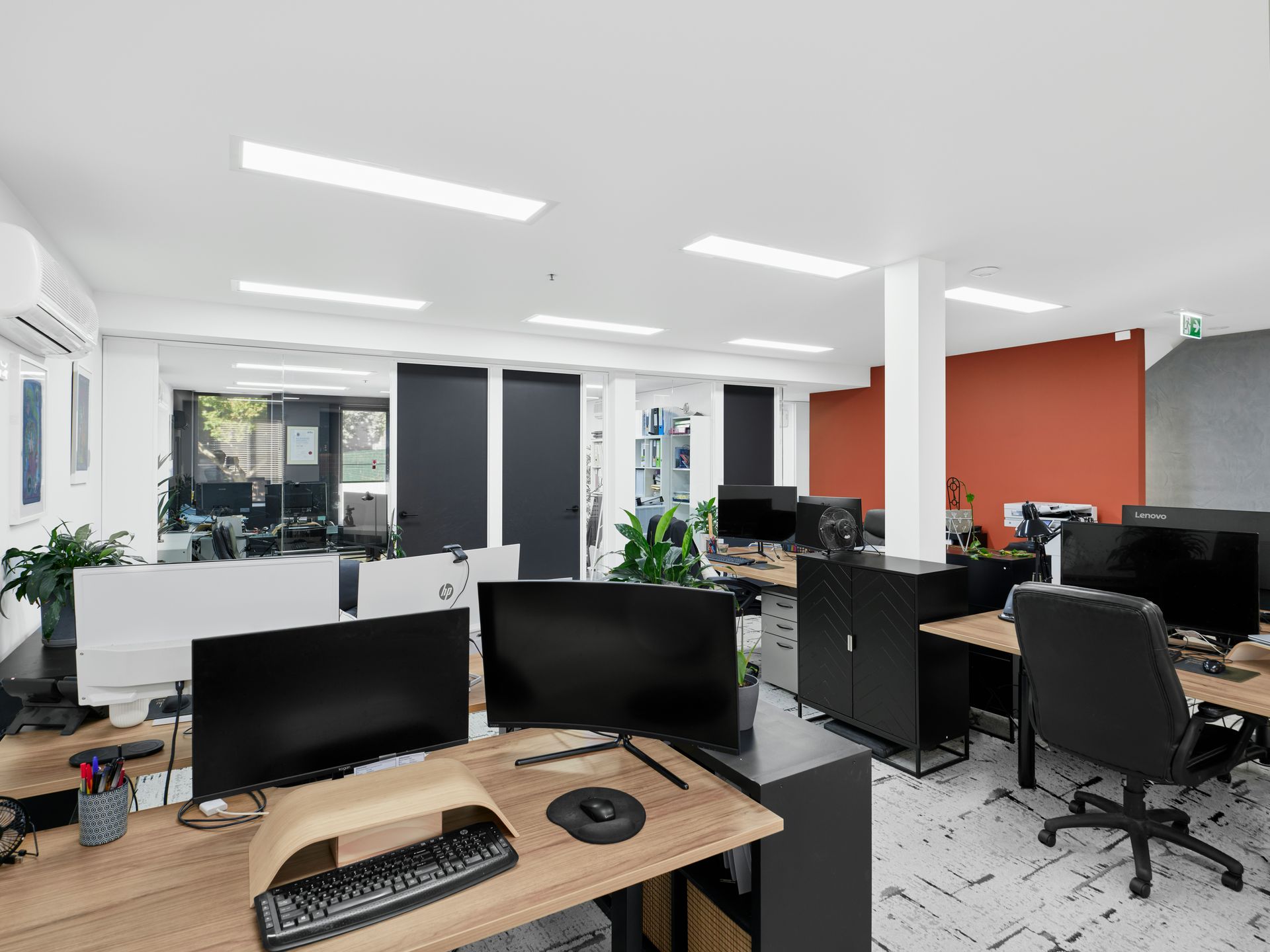 Office workspace with multiple desks, computer monitors, and chairs. Bright overhead lighting.