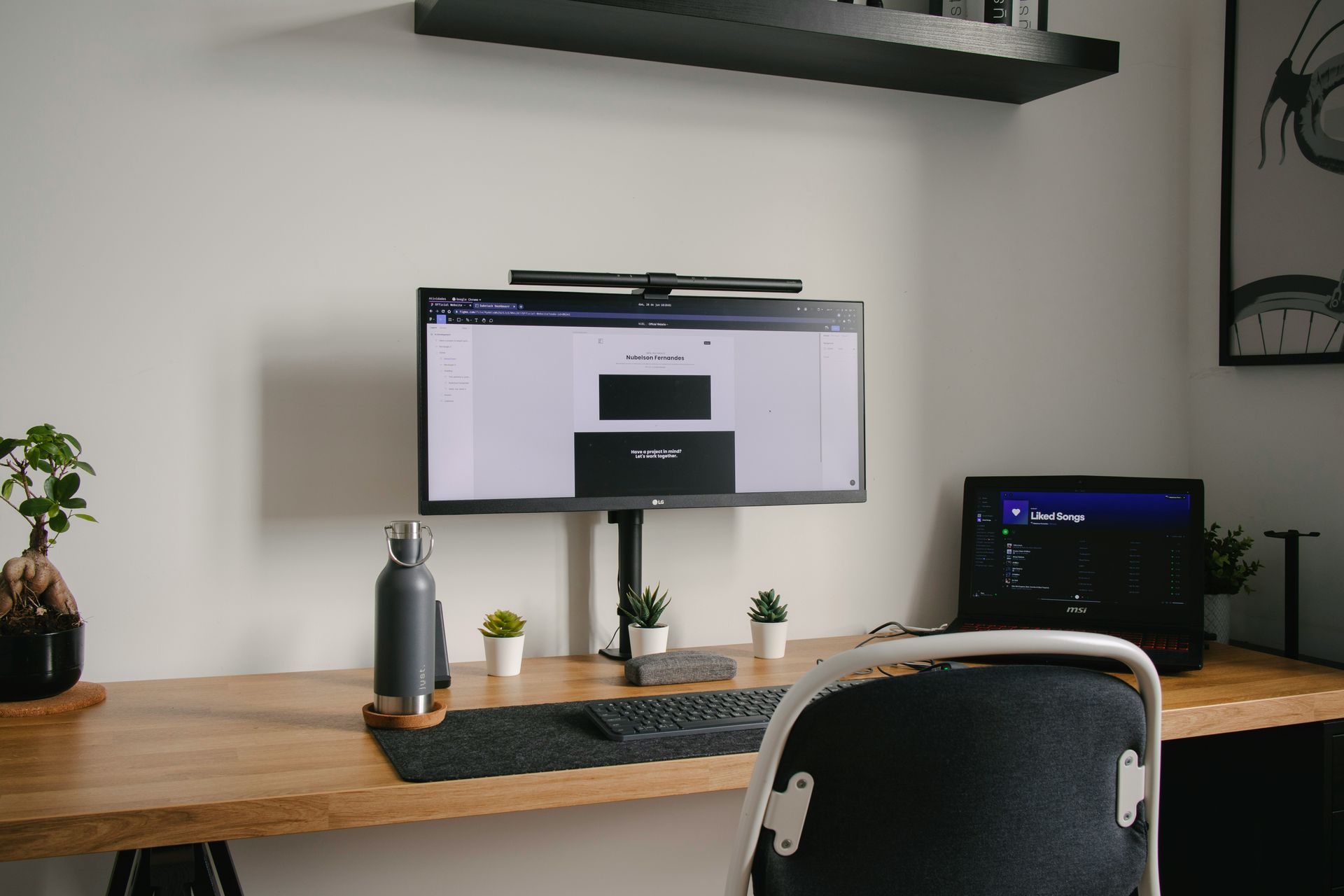 Workspace with monitor, keyboard, laptop, plants, and water bottle on a wooden desk against a white wall.