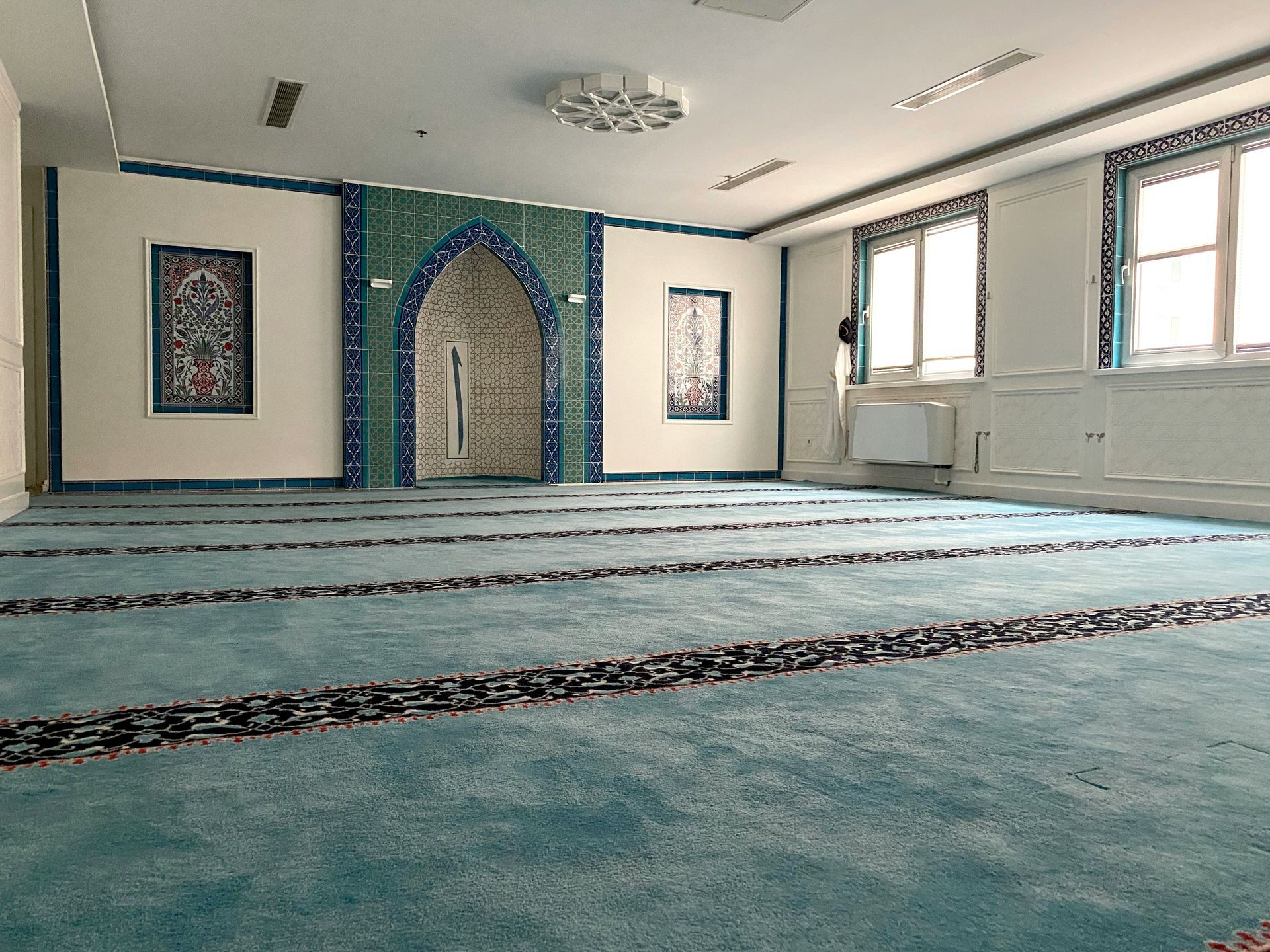 Interior view of a mosque prayer hall with blue carpet and tiled archway. Windows let in light.