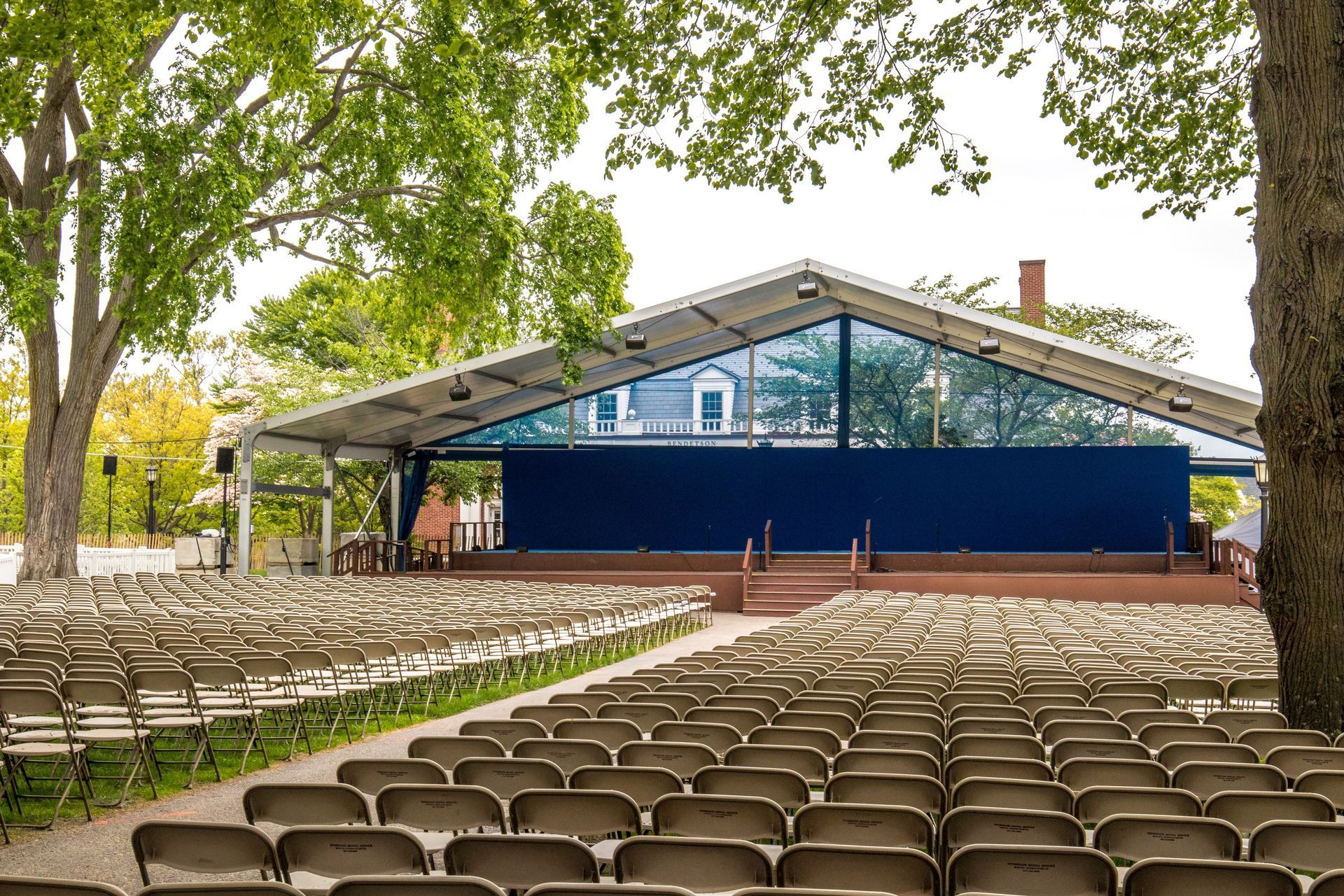 Outdoor theater with rows of empty chairs facing a blue stage beneath a peaked roof. Trees frame the scene.