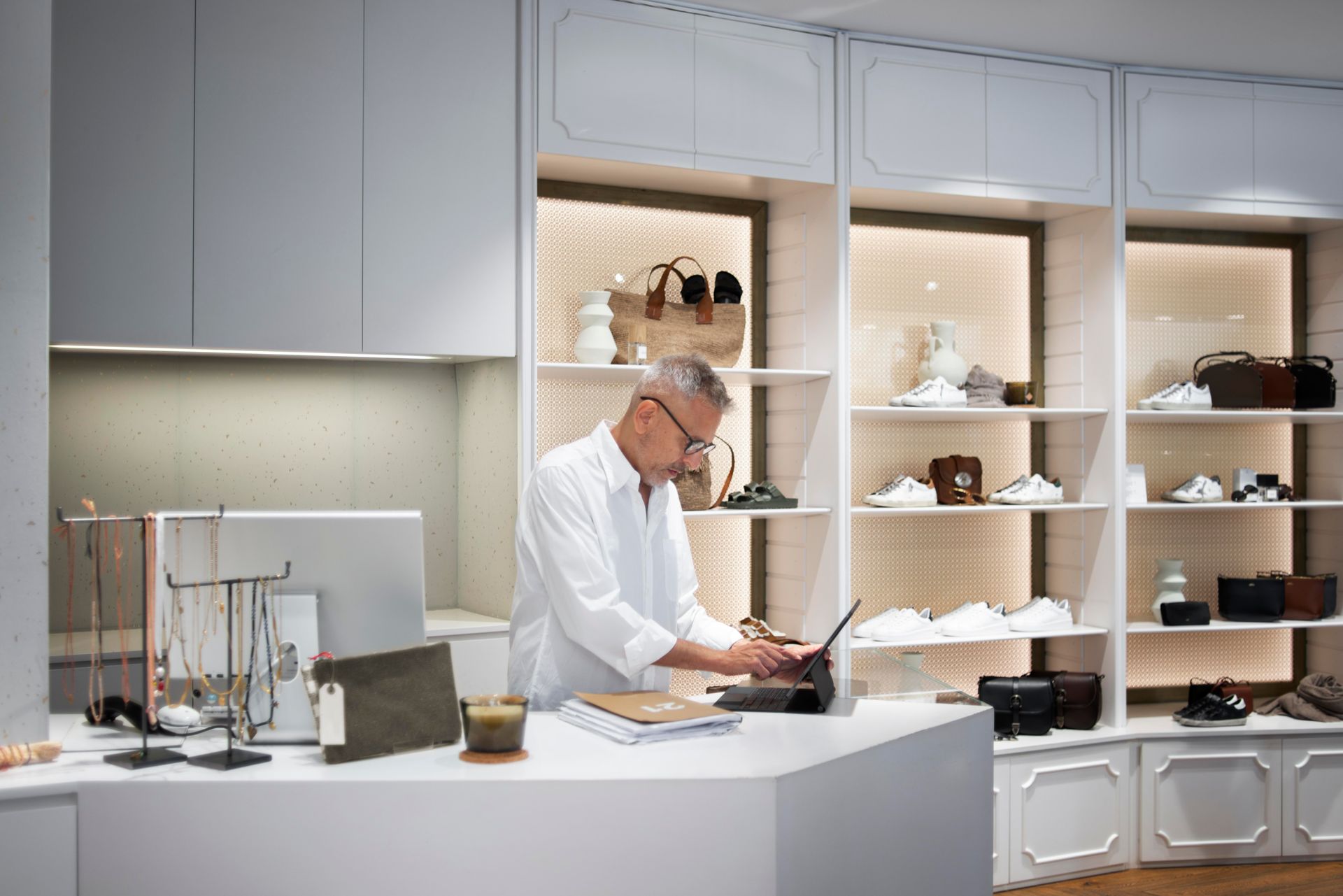 Man in white shirt uses a laptop at a retail counter, with shelves of shoes and bags in the background.