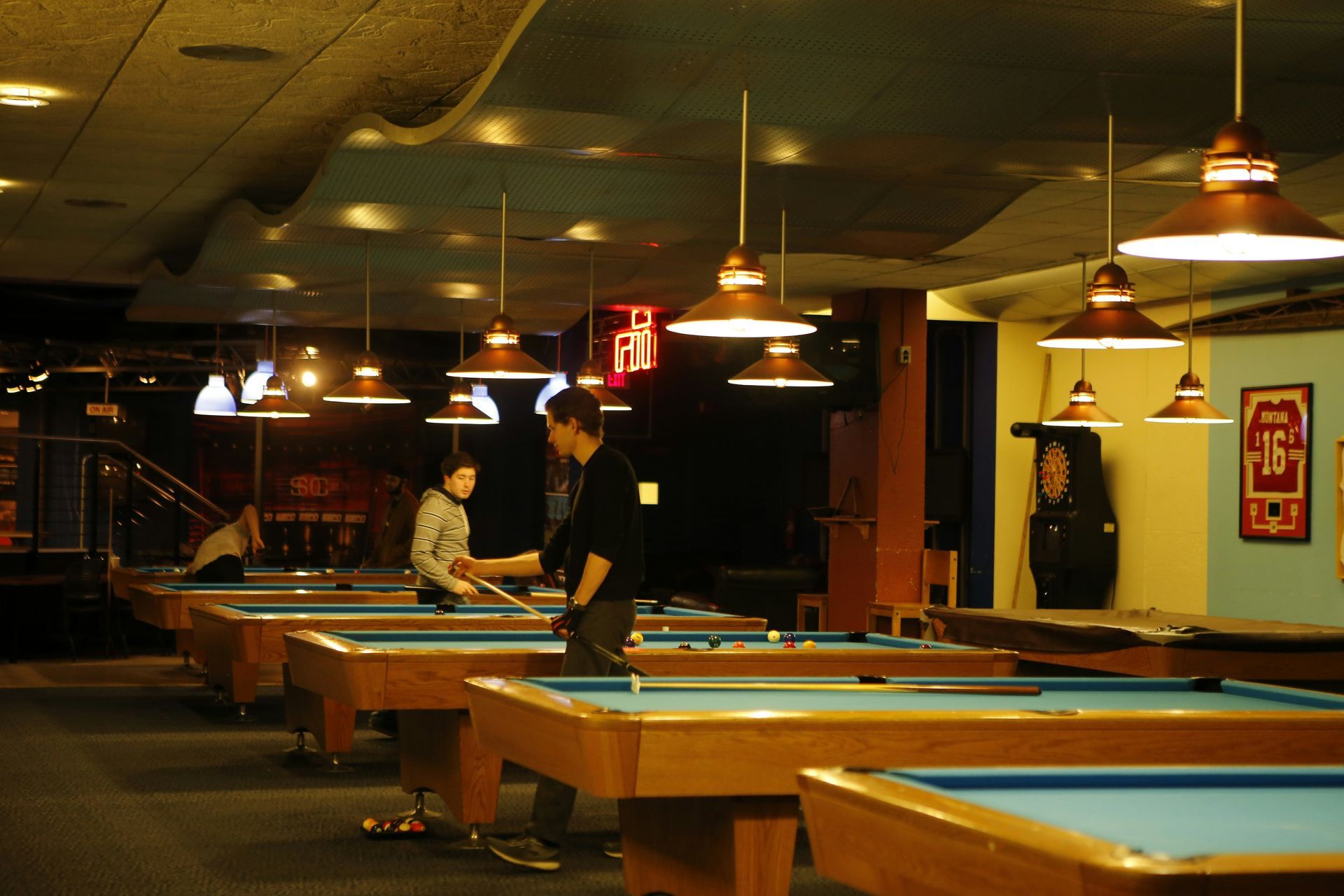 Pool hall with multiple tables, overhead lights, and two people playing pool.