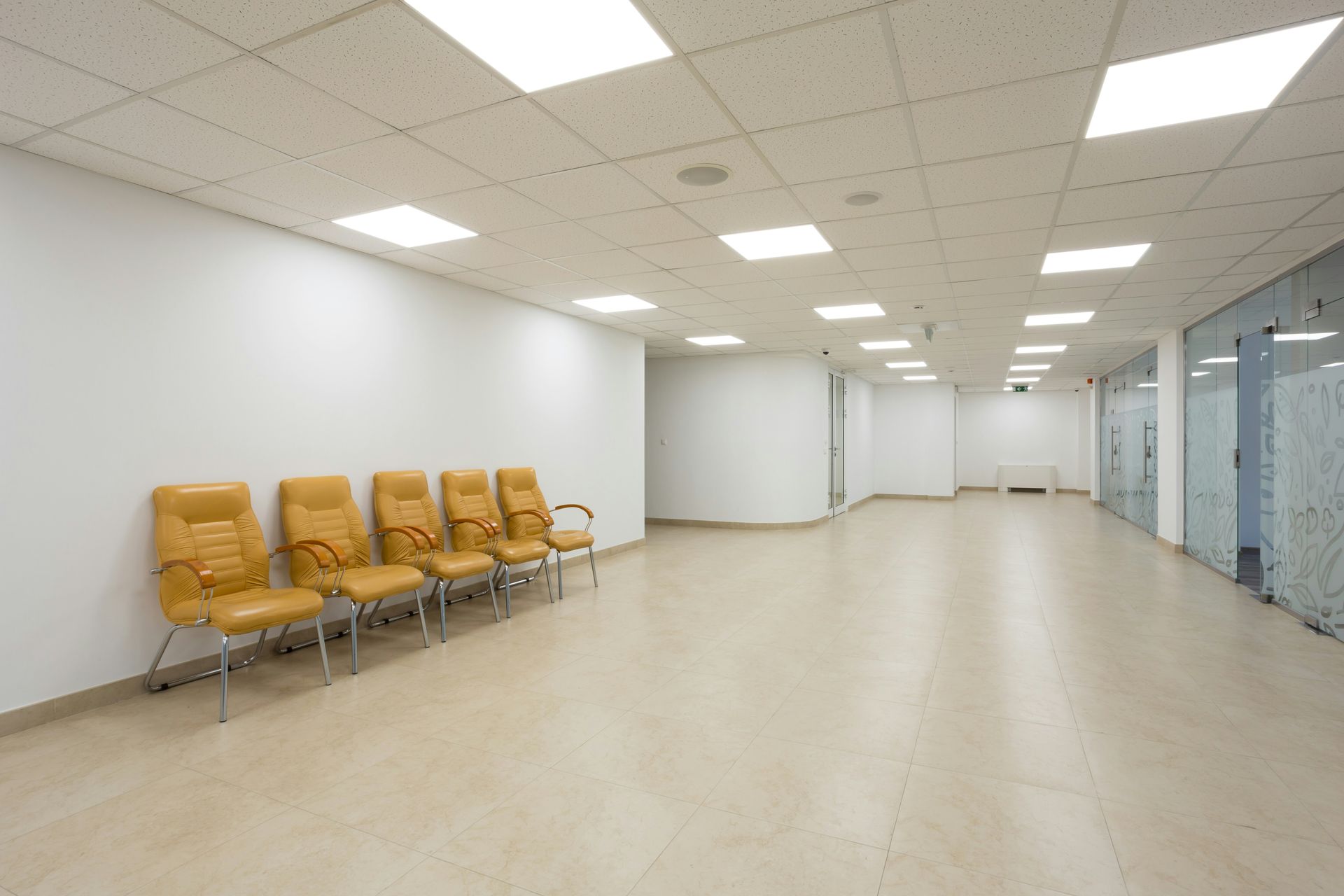 Empty waiting room with tan chairs against a white wall and a glass-walled corridor on the right.