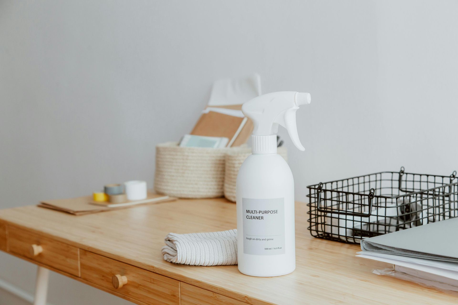 White spray bottle on a wooden desk with a cloth, basket, and wire bin.