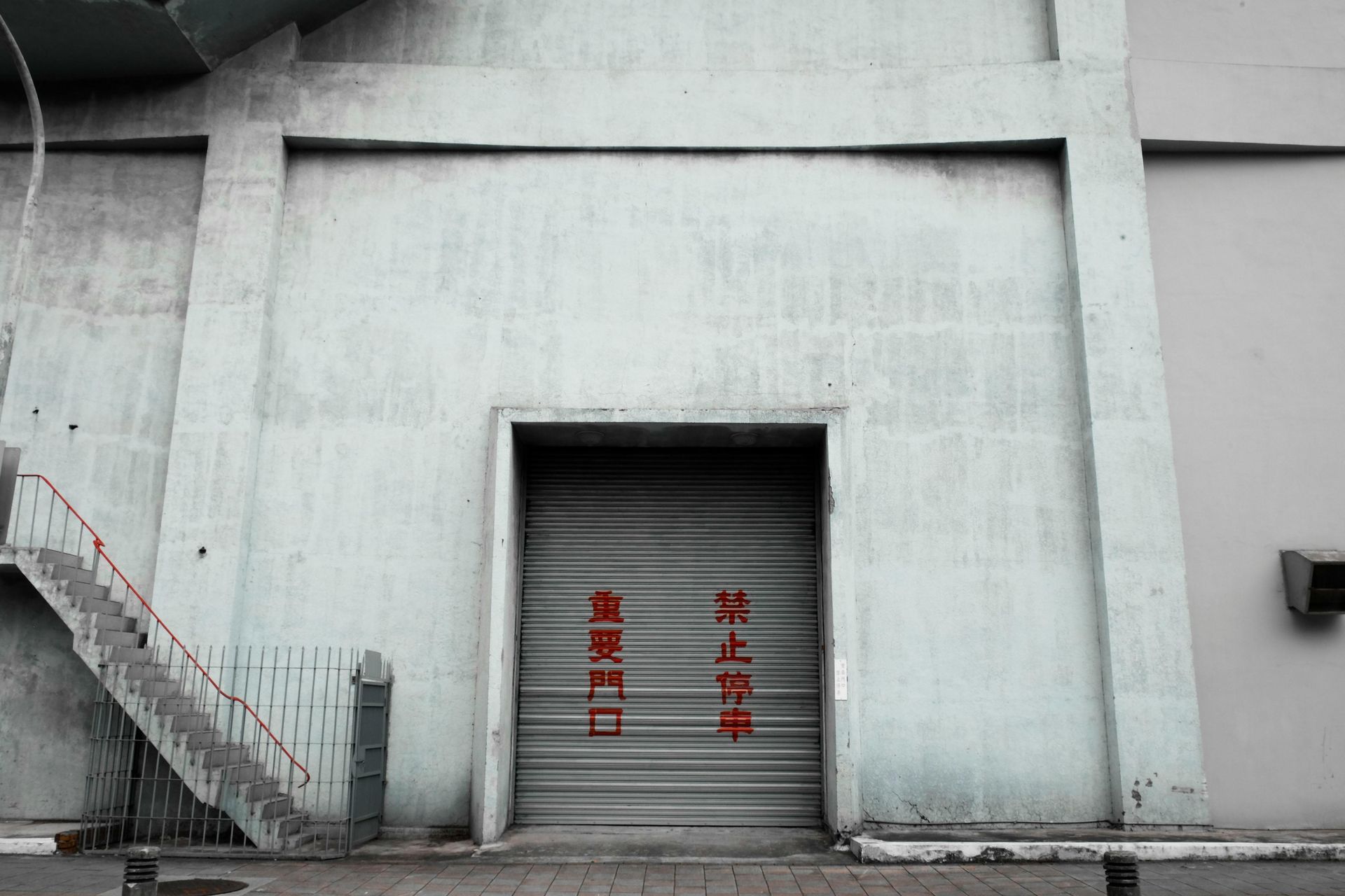 Concrete industrial building with a closed garage door. Red text on the door reads in a foreign language. Stairs on the left.