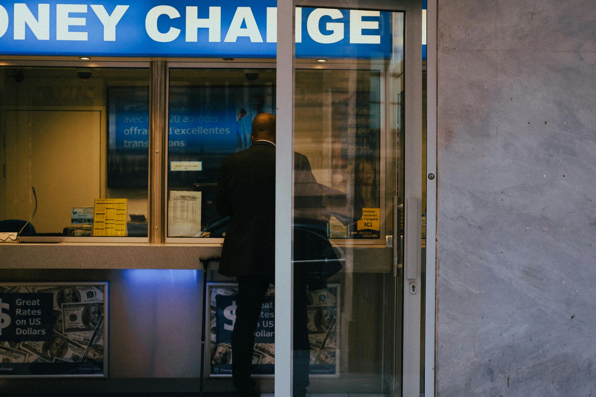 Person at a money exchange booth, looking at the teller behind glass doors. Blue neon sign.