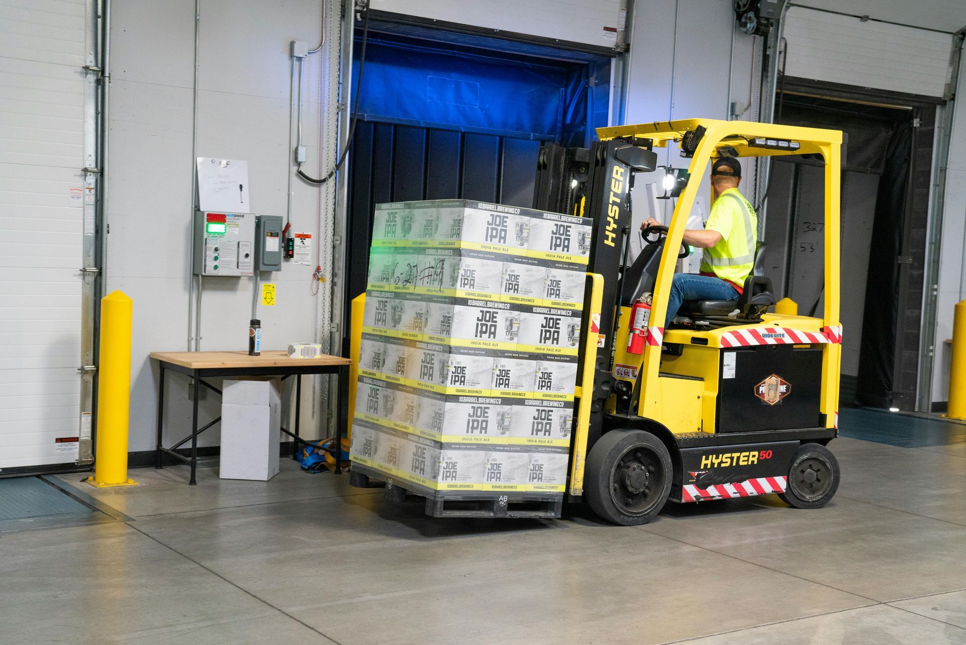 Forklift carrying stacked boxes at a loading dock; worker in safety vest; green light visible.