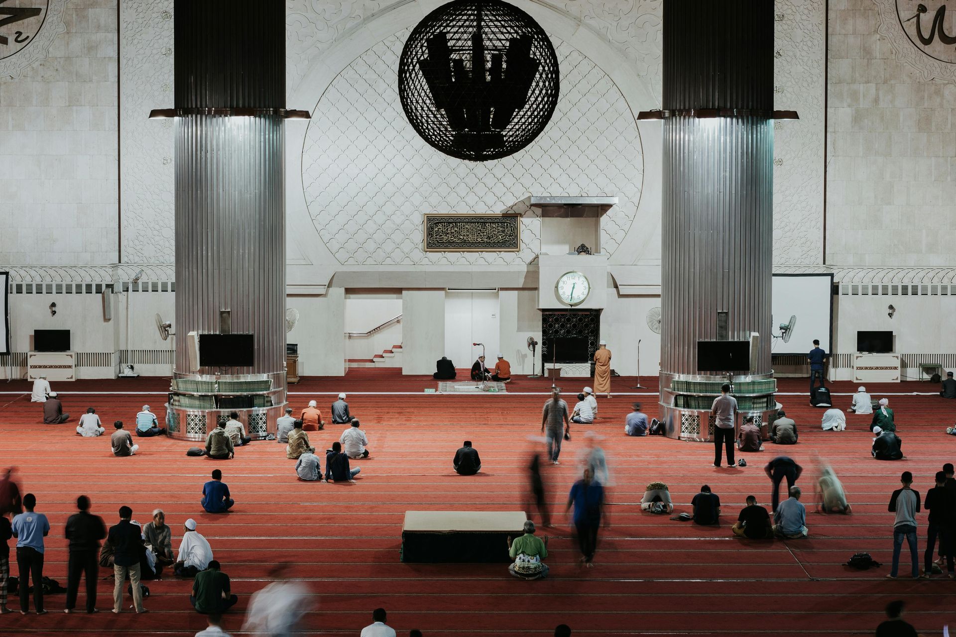 People praying inside a mosque with red carpets, large columns, and a clock on the wall.