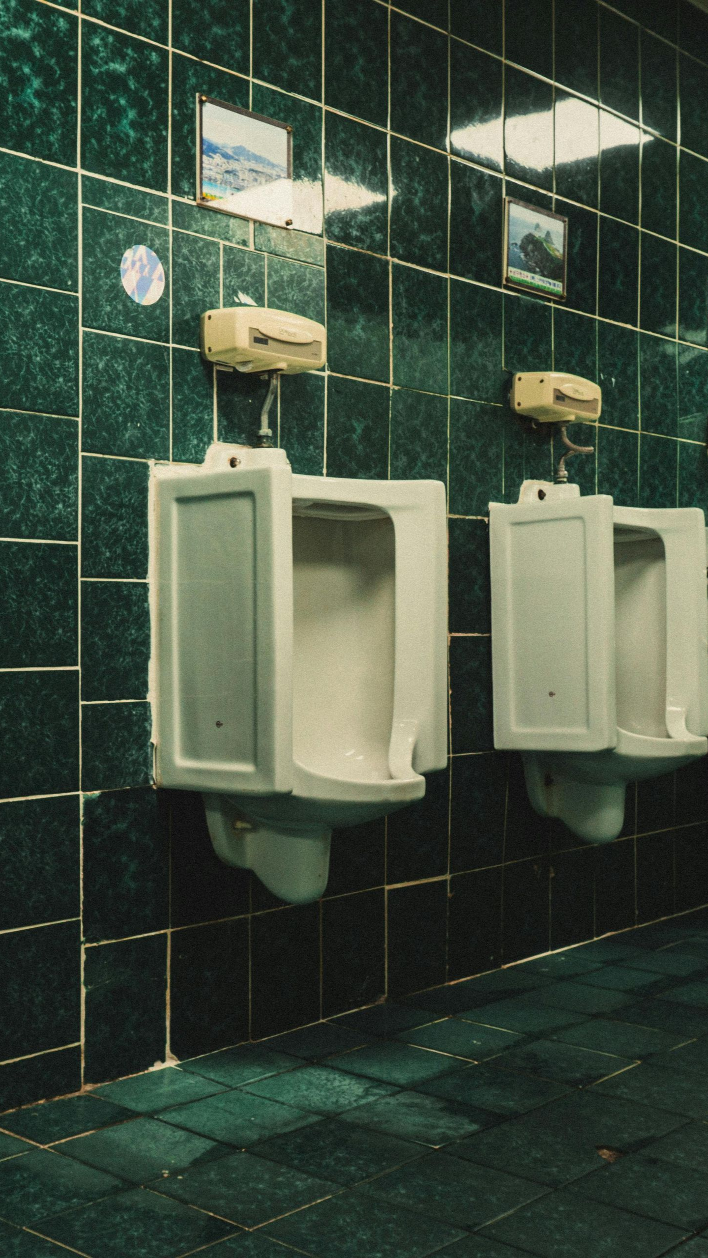 Two white urinals with cream-colored automatic flushers on a green tiled wall in a restroom.