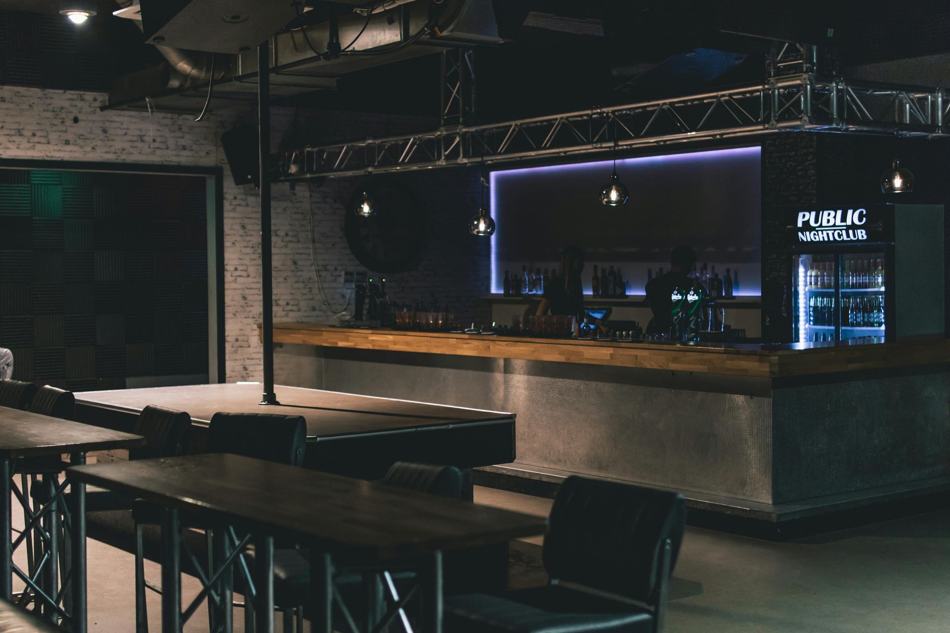 Interior view of a modern bar with a long wooden counter and seating. Dim lighting, brick and metal details.
