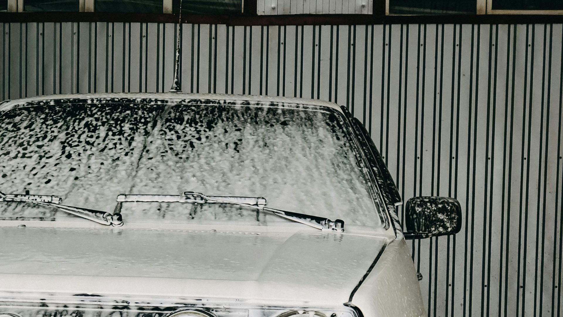 White car covered in thick foam at a car wash, against a corrugated metal wall.